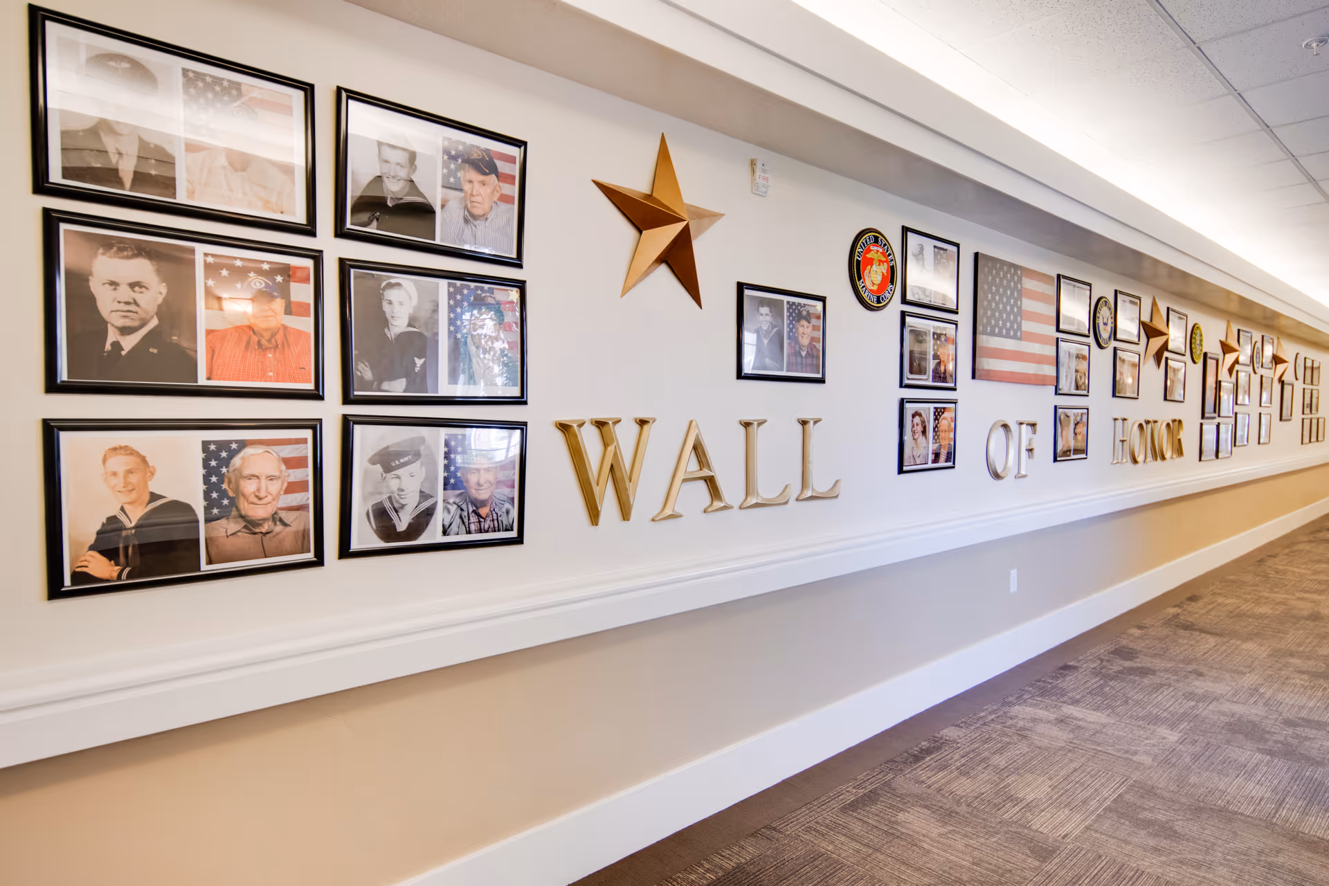 A bright hallway interior displaying a long 'Wall of Honor' with framed photos, American flags, decorative stars and large gold letters.