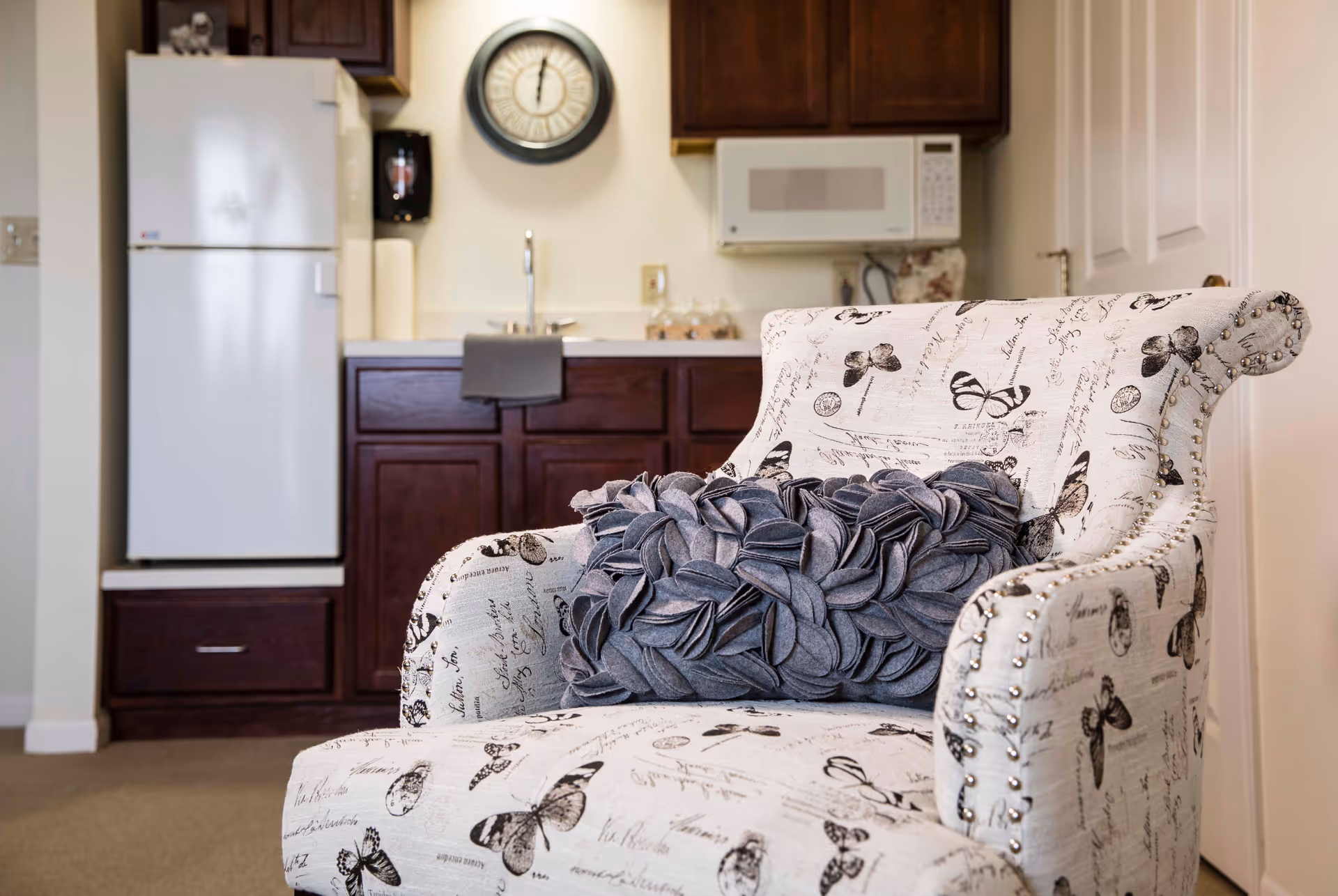 A cozy interior scene featuring a white armchair with butterfly and script patterns, adorned with a textured gray pillow. Behind the chair is a small kitchen area with dark wooden cabinets, a white refrigerator, a microwave, a sink, and a wall clock.