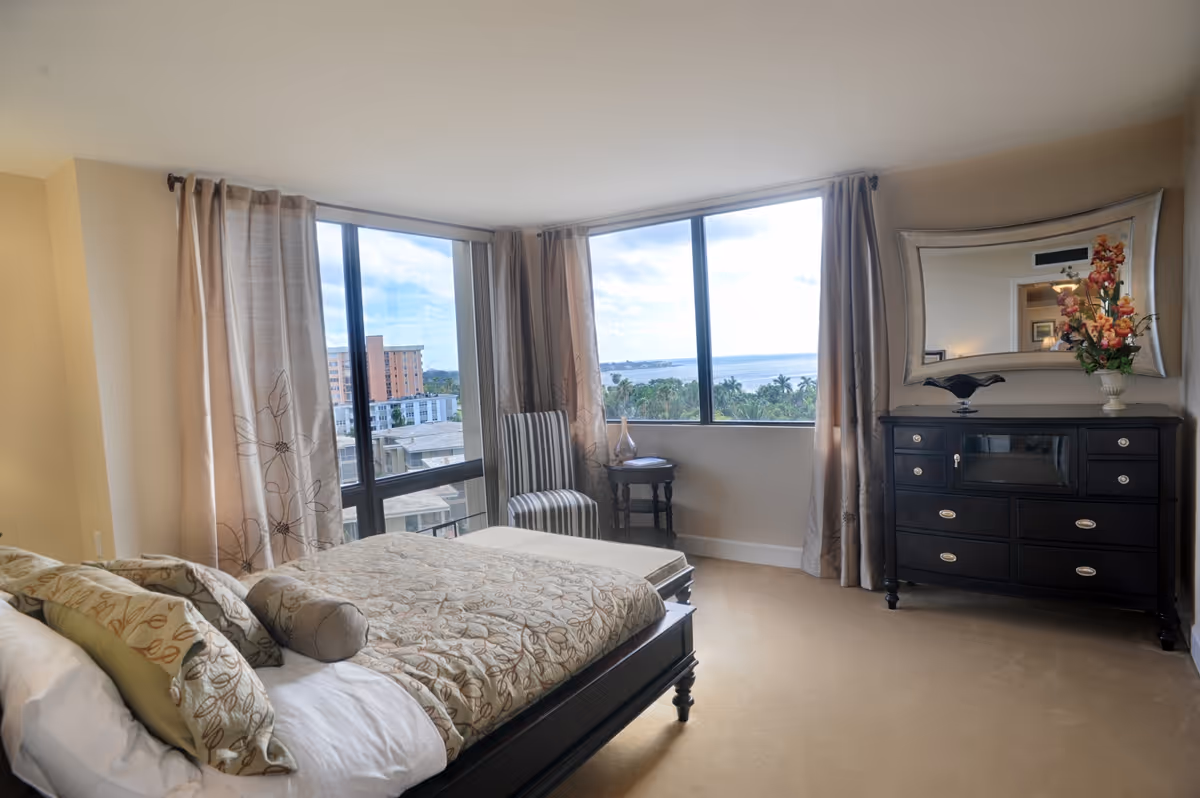 A bright bedroom with large windows offering a view of buildings, palm trees, and water. The room features a bed with patterned bedding and multiple pillows, a striped chair next to a small round table, and a dark wooden dresser with a large decorative mirror above it. A vase with flowers and a decorative bowl are placed on the dresser.