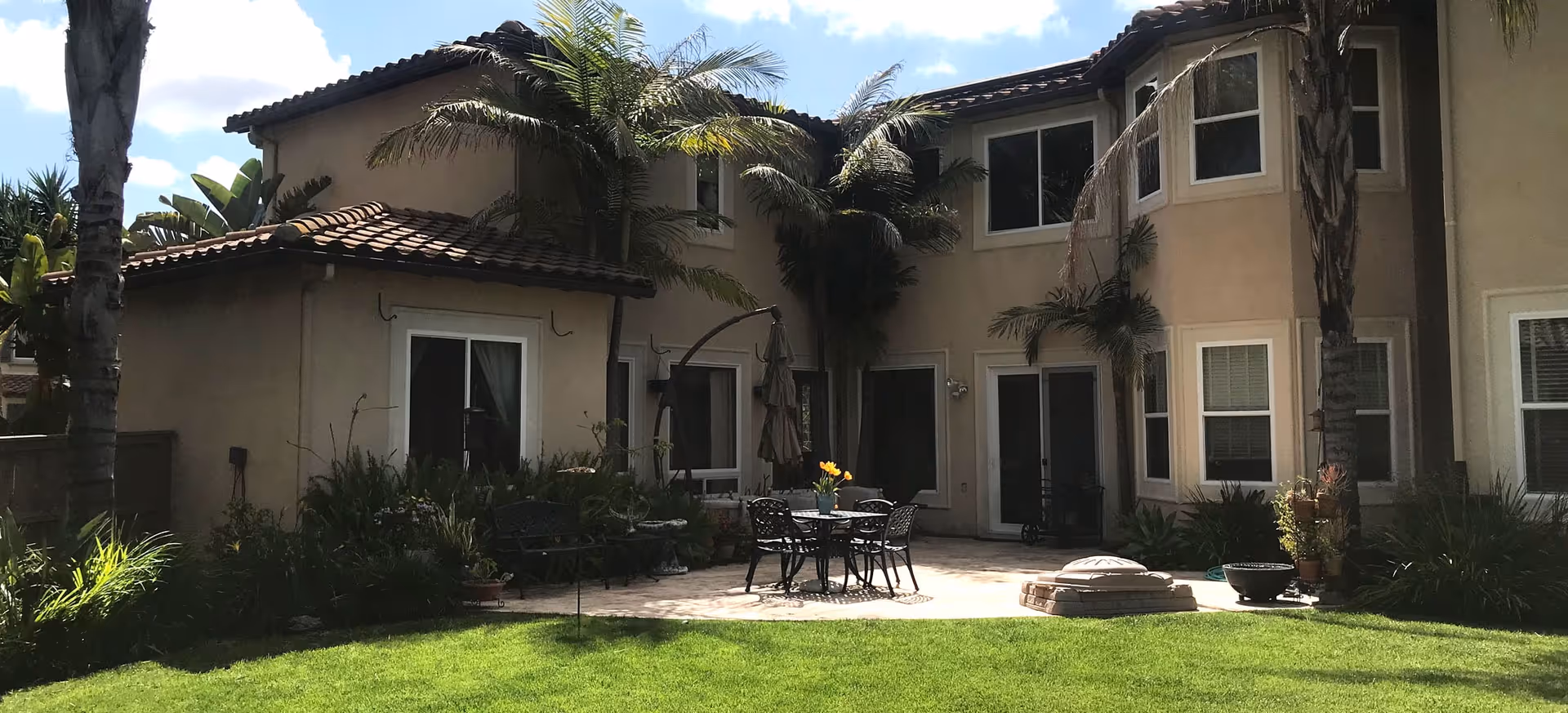 Outdoor patio area of a residential building with beige walls and tiled roof. The patio has a round table with four chairs and an umbrella, surrounded by palm trees and green grass lawn.