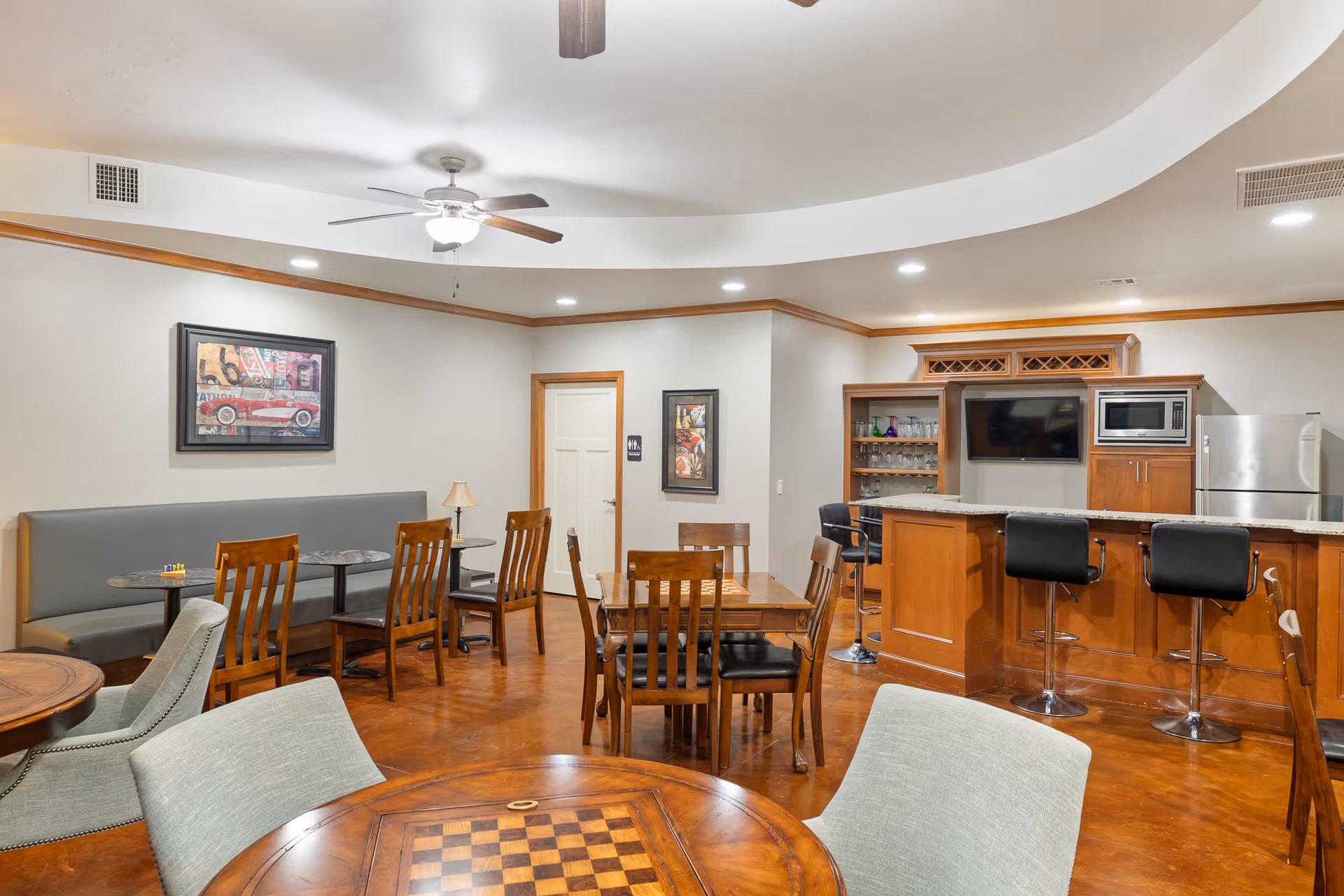 A spacious common area with multiple wooden tables and chairs, a built-in bench along one wall, a ceiling fan, and a kitchenette with a granite countertop, bar stools, a microwave, and a refrigerator. The room has warm lighting, framed artwork on the walls, and a polished wooden floor.