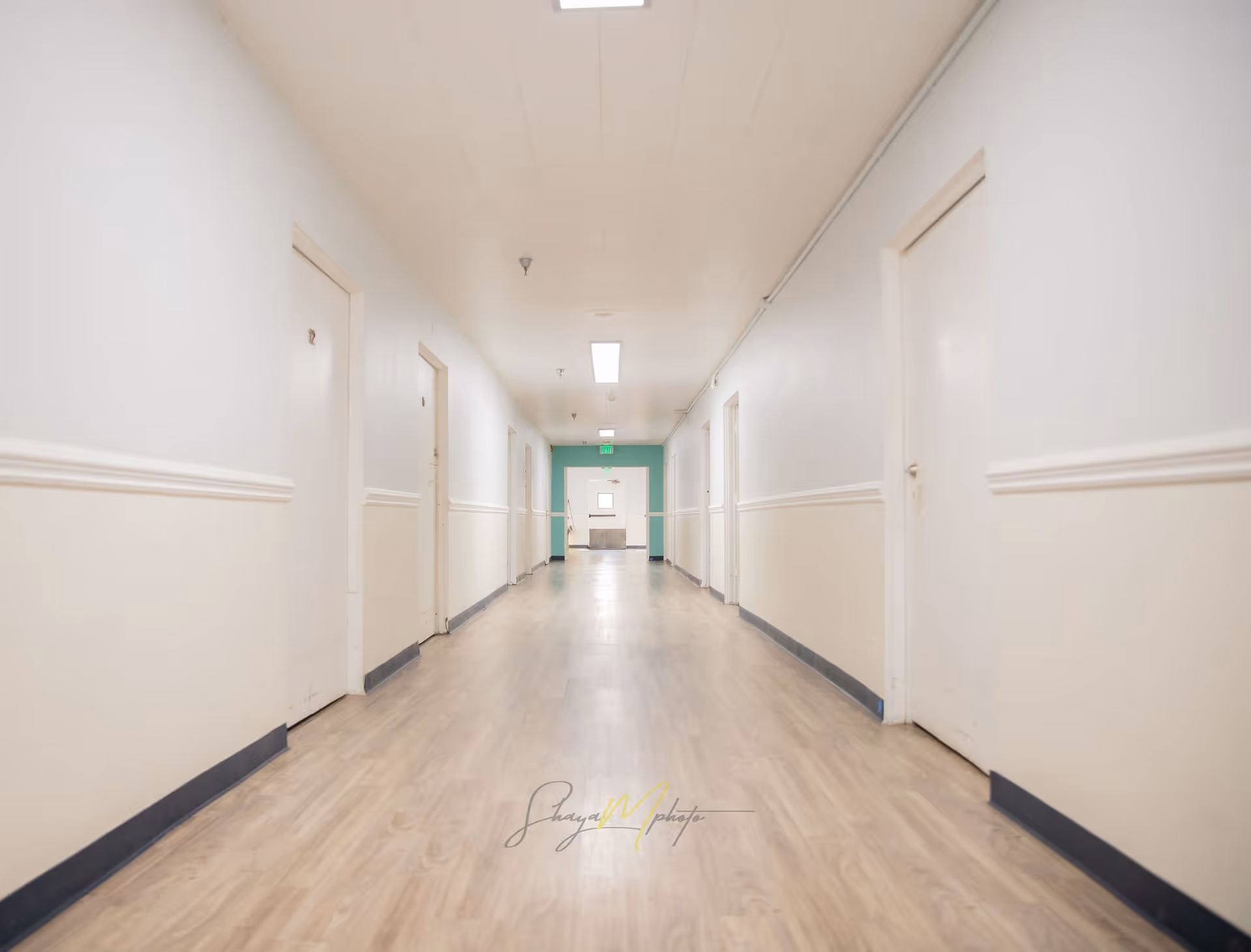 A long, empty hallway in an assisted living facility with light-colored walls, multiple closed white doors on both sides, and a light wood floor. The hallway is well-lit with ceiling lights and has a green exit sign at the far end.