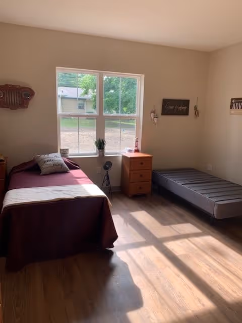 A bright bedroom with two beds, one with a maroon bedspread and a pillow, and the other a bare mattress. There is a wooden nightstand between the beds with a small plant and a decorative item on it. A large window lets in natural light, casting shadows on the wooden floor. The walls are decorated with small wall hangings and signs.