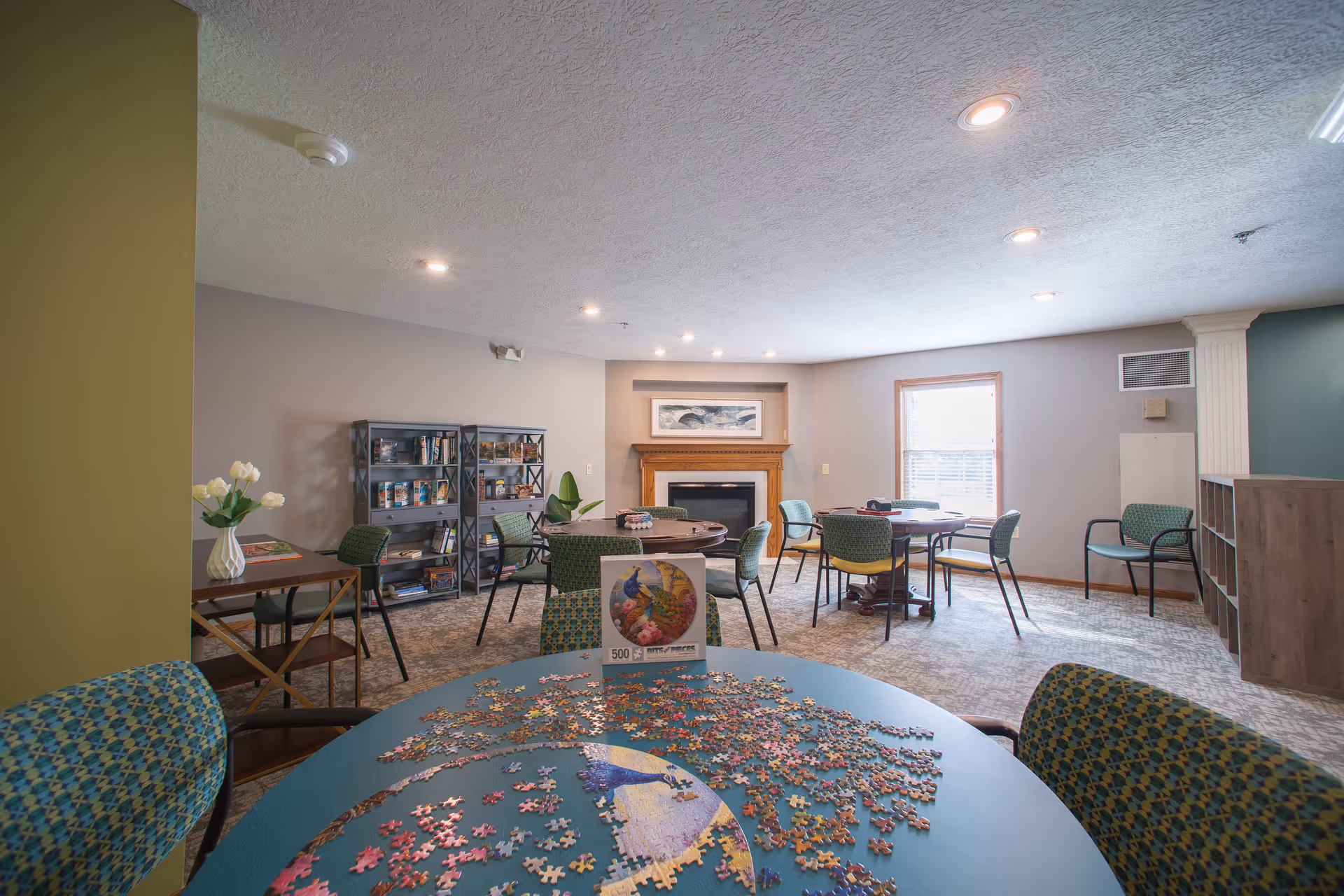 A well-lit common room with several tables and chairs arranged for group activities. One table in the foreground has a partially completed jigsaw puzzle with a colorful bird image. The room features a fireplace with a framed picture above it, a bookshelf filled with books and games, and large windows letting in natural light. The walls are painted in neutral tones, and the carpet has a subtle pattern.