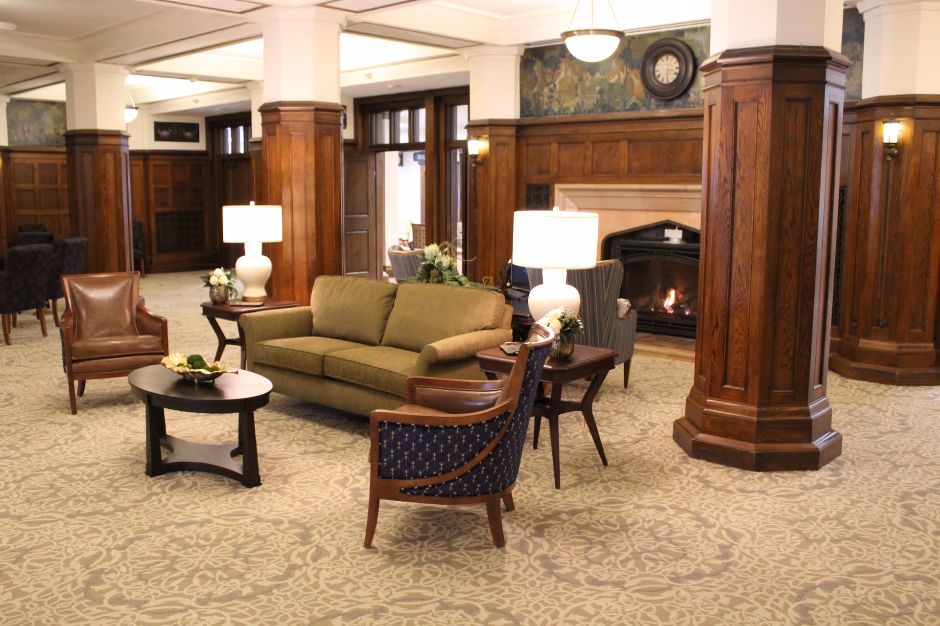 A cozy living room area with a green sofa, two armchairs, a coffee table, and side tables with lamps. The room features wooden columns, a fireplace with a fire burning, and patterned carpet flooring.