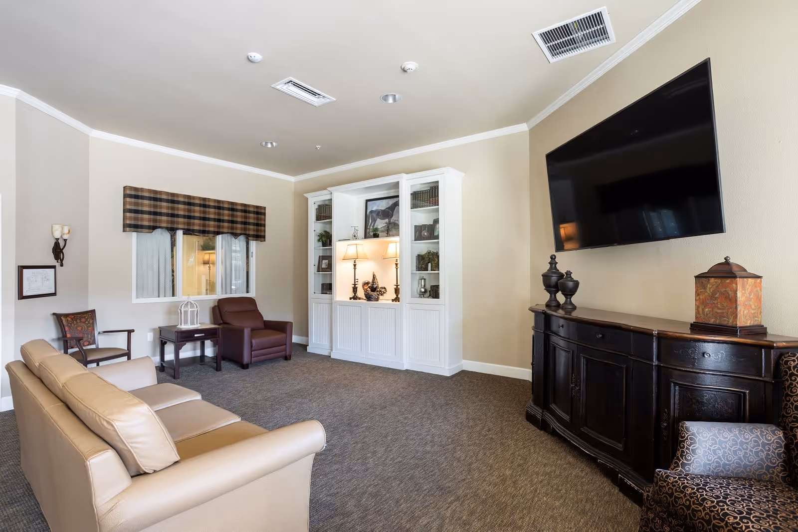 A cozy living room with a beige leather sofa, a brown armchair, a wooden side table, and a white built-in cabinet with lamps and decorative items. A large flat-screen TV is mounted on the wall above a dark wooden console table. The room has beige walls, carpeted floor, and a window with a plaid valance.