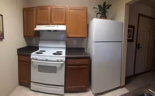 A small kitchen area featuring a white electric stove with four burners and an oven, wooden cabinets above and below the countertop, a white refrigerator with a green potted plant on top, and a doorway leading to another room.