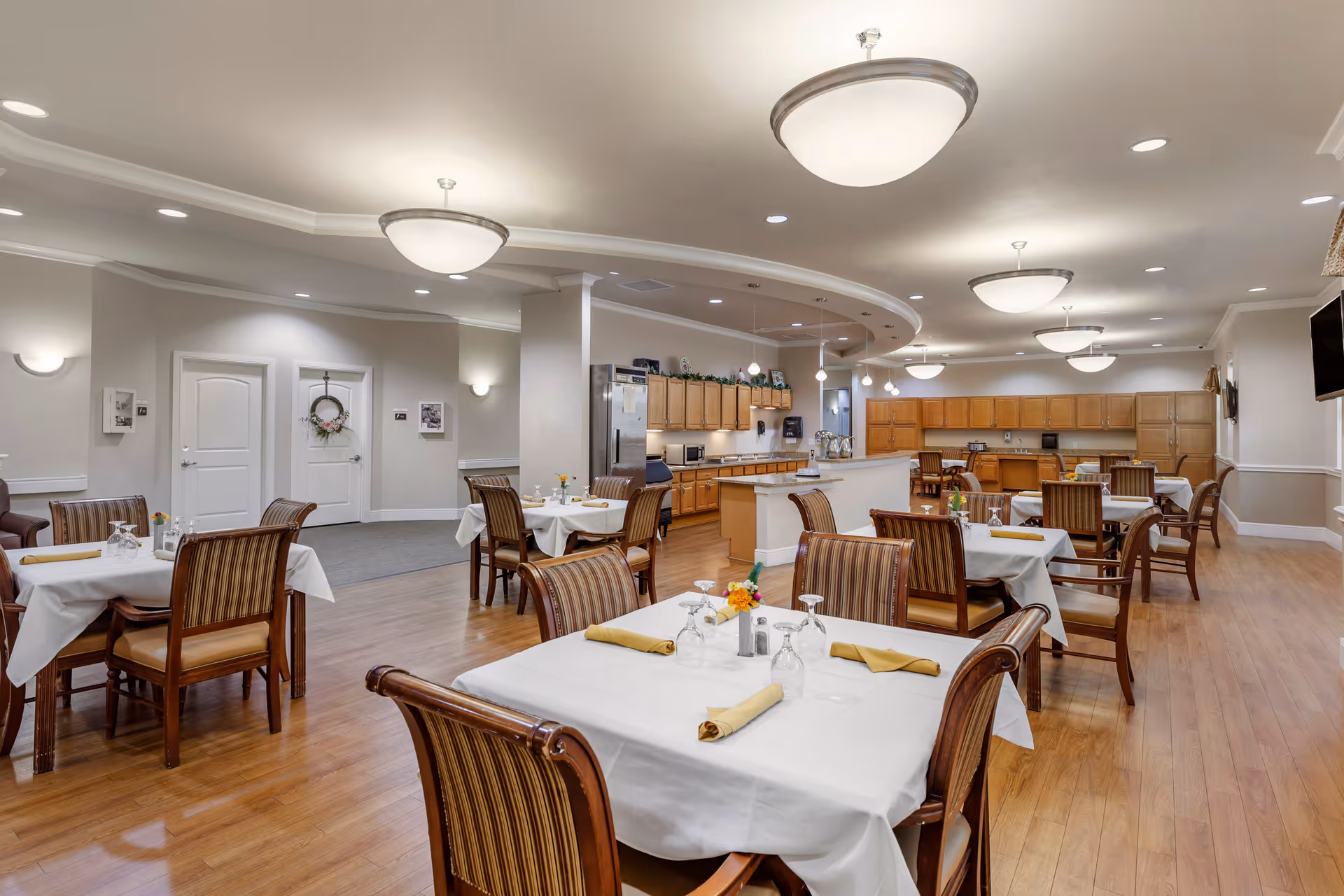 A spacious dining room with multiple tables covered in white tablecloths, each set with glasses, napkins, and small flower arrangements. The room features wooden chairs with striped cushions, wooden flooring, and a kitchen area with wooden cabinets and stainless steel appliances in the background. The ceiling has recessed lighting and several large, round light fixtures.
