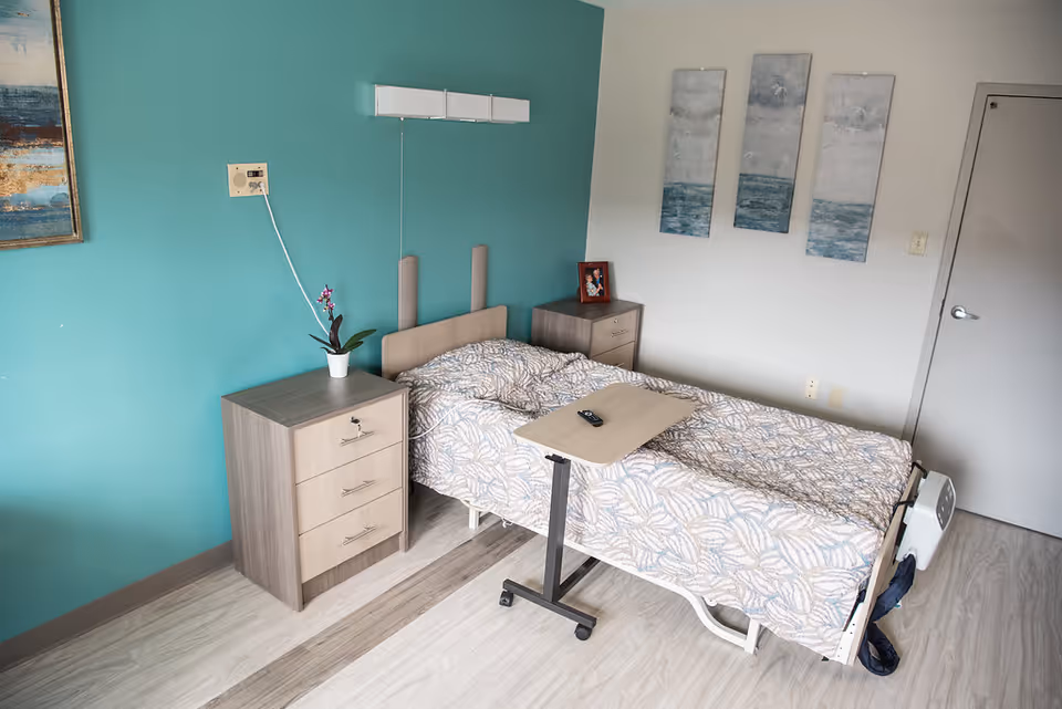 A single bed in a nursing care facility room with a patterned bedspread, an adjustable overbed table with a remote control on it, two bedside cabinets with a small plant and a framed photo, teal and white walls with three ocean-themed paintings, and a closed door.