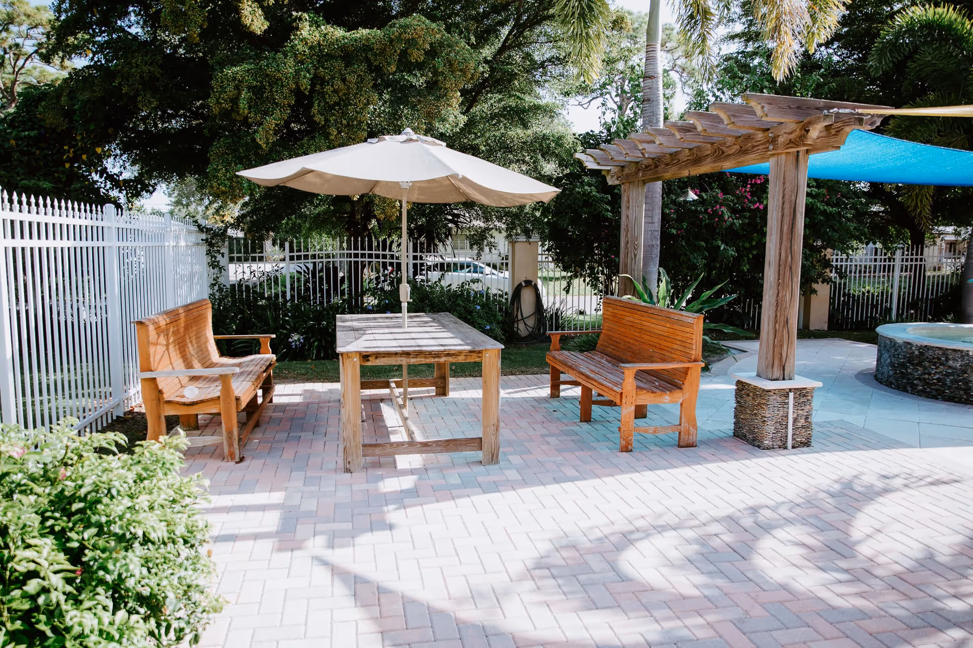 Outdoor patio area with two wooden benches facing a wooden table with a large beige umbrella. The area is paved with light-colored bricks and surrounded by greenery, including trees and bushes. There is a wooden pergola with a blue shade cloth and a circular stone water feature or hot tub to the right.