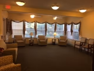 A spacious living room with beige walls and carpeted floor, featuring a row of large windows with valance curtains. The room is furnished with several beige armchairs arranged around small tables with lamps, and additional wooden chairs lined up against the wall.