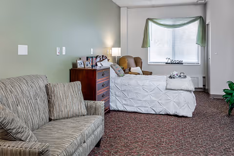 A cozy assisted living room featuring a patterned sofa with two cushions on the left, a wooden chest of drawers with framed photos and books, a single bed with a white textured bedspread, a brown armchair, and a window with green curtains and a decorative 'Dream' sign on the windowsill.
