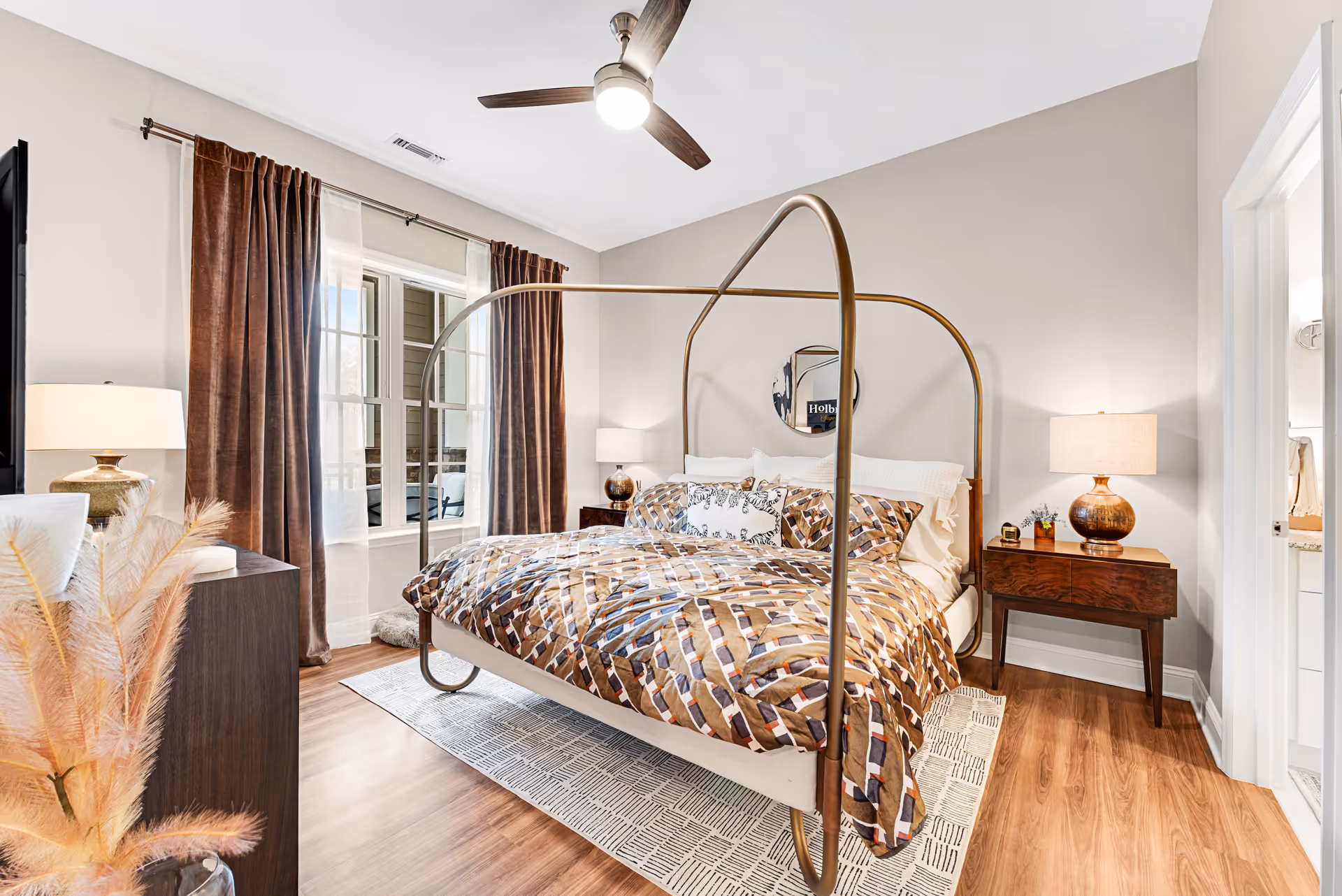 A bright bedroom with a modern four-poster bed featuring a geometric patterned comforter. The room has wooden flooring, a ceiling fan with light, two bedside tables with lamps, and a window with brown curtains and sheer white drapes. A round mirror hangs on the wall above the bed, and a doorway leads to an adjacent bathroom.