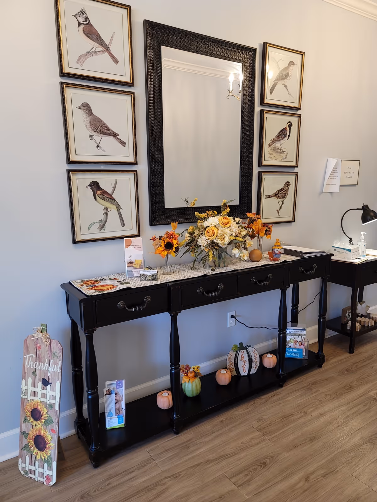 Decorative entry console table under a large mirror with framed bird prints, a floral arrangement and seasonal pumpkins on display.