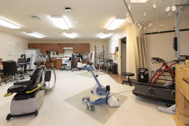 A rehabilitation room in a senior living facility featuring exercise equipment including a stationary bike, a treadmill, and a walker. The room has a kitchenette area with cabinets, a refrigerator, and a stove in the background. There are chairs and various medical or therapy devices around the room. Snowflake decorations hang from the ceiling.