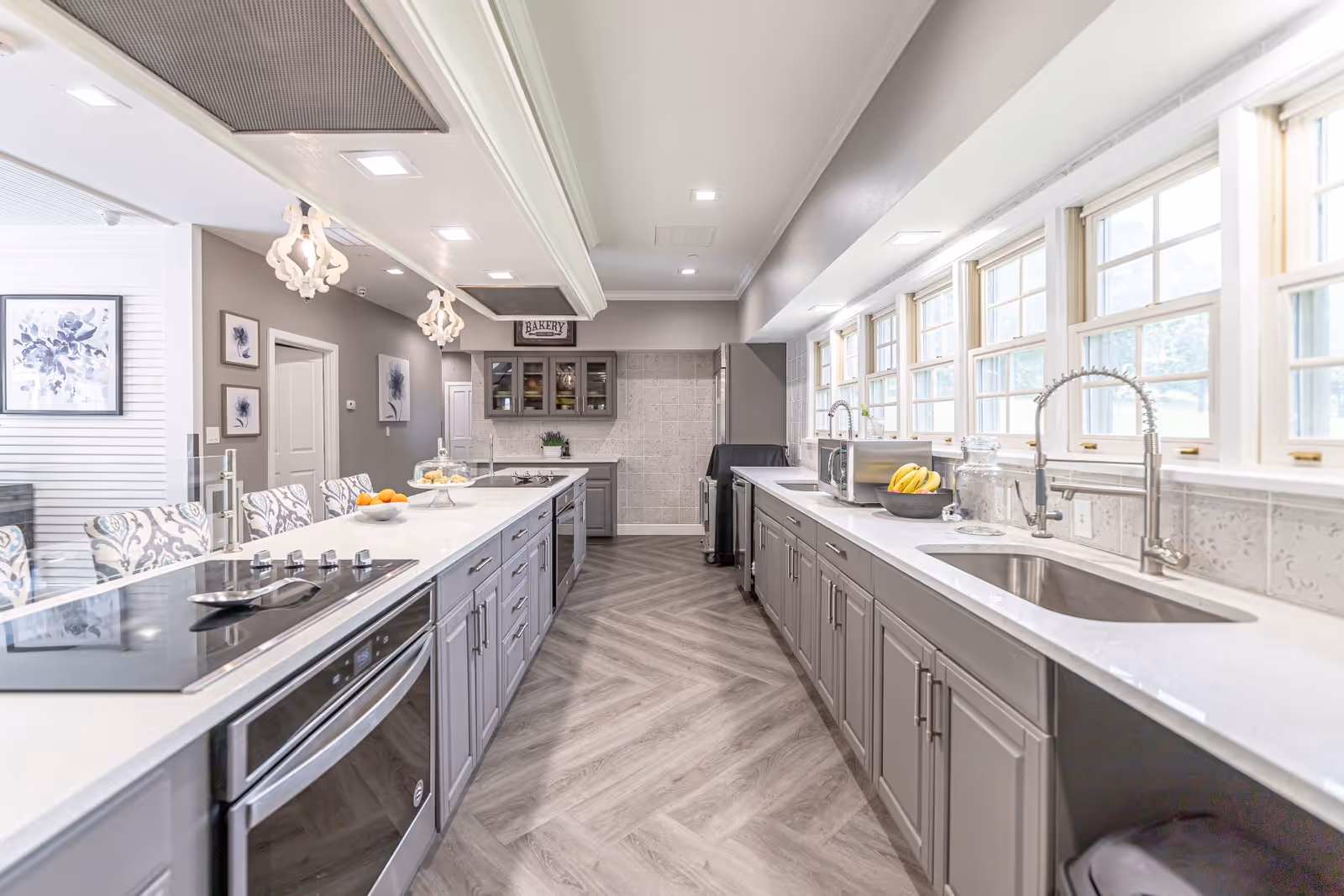 A modern kitchen with gray cabinets and white countertops on both sides. The left side features a stovetop and oven with bar stools along the counter, while the right side has a sink, microwave, and a bowl of bananas. Multiple windows above the right counter let in natural light. The floor has a herringbone pattern, and decorative light fixtures hang from the ceiling. Artwork and a sign reading 'Bakery' are visible on the far wall.