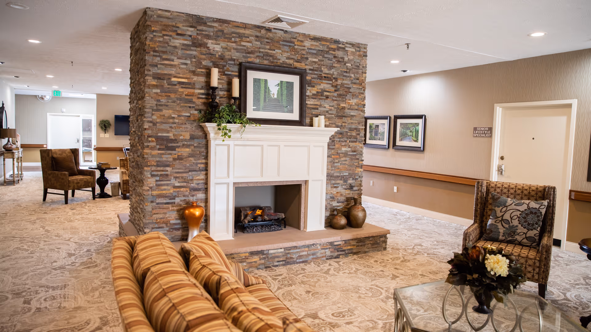A cozy senior living facility common area with a stone fireplace in the center, decorated with candles, vases, and a framed picture above the mantel. The room features patterned carpet, a striped sofa, an armchair with a floral cushion, a glass coffee table with a flower arrangement, and framed artwork on the walls.