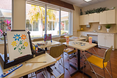 Bright activity room with tables and chairs set up for crafts and games, an easel with a painting, a kitchenette with cabinets, and large windows overlooking a courtyard.