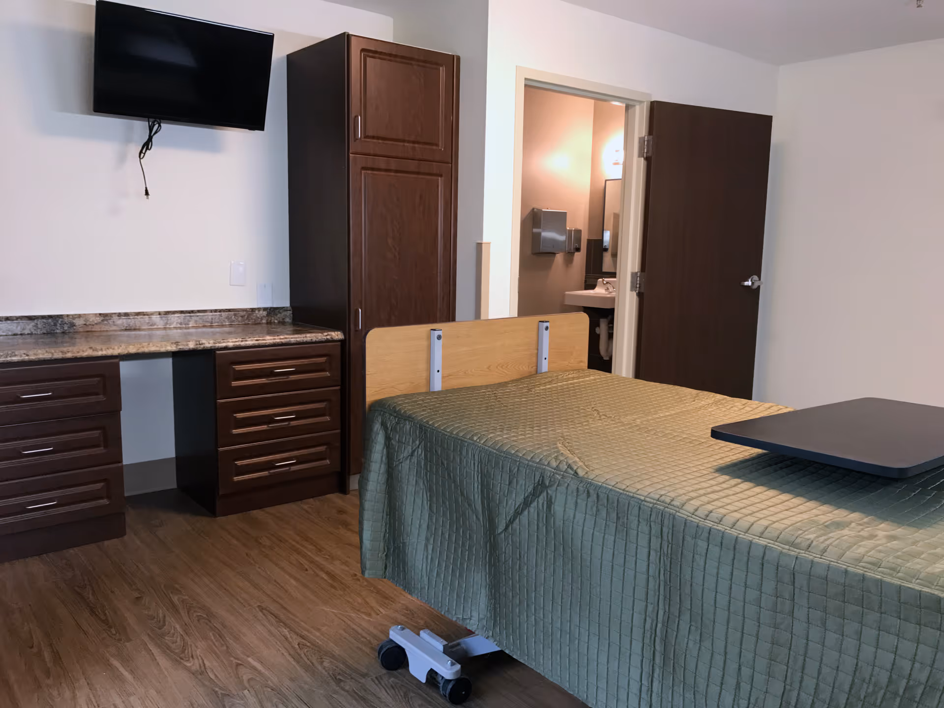 A senior living facility room with a bed covered in a green quilted blanket, a wooden headboard, and a black overbed table. The room has wooden flooring, dark wood cabinets and drawers, a granite countertop, a wall-mounted flat-screen TV, and an open door leading to a bathroom with a sink and paper towel dispenser.