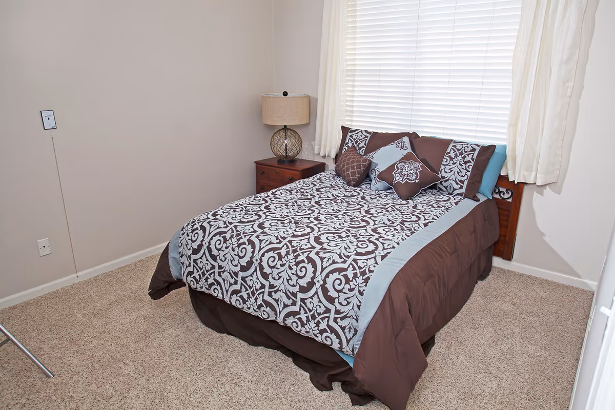 A bedroom with a bed covered in a brown and light blue patterned comforter and matching pillows. There is a wooden nightstand with a lamp next to the bed, beige carpet flooring, and a window with white blinds and curtains behind the bed.