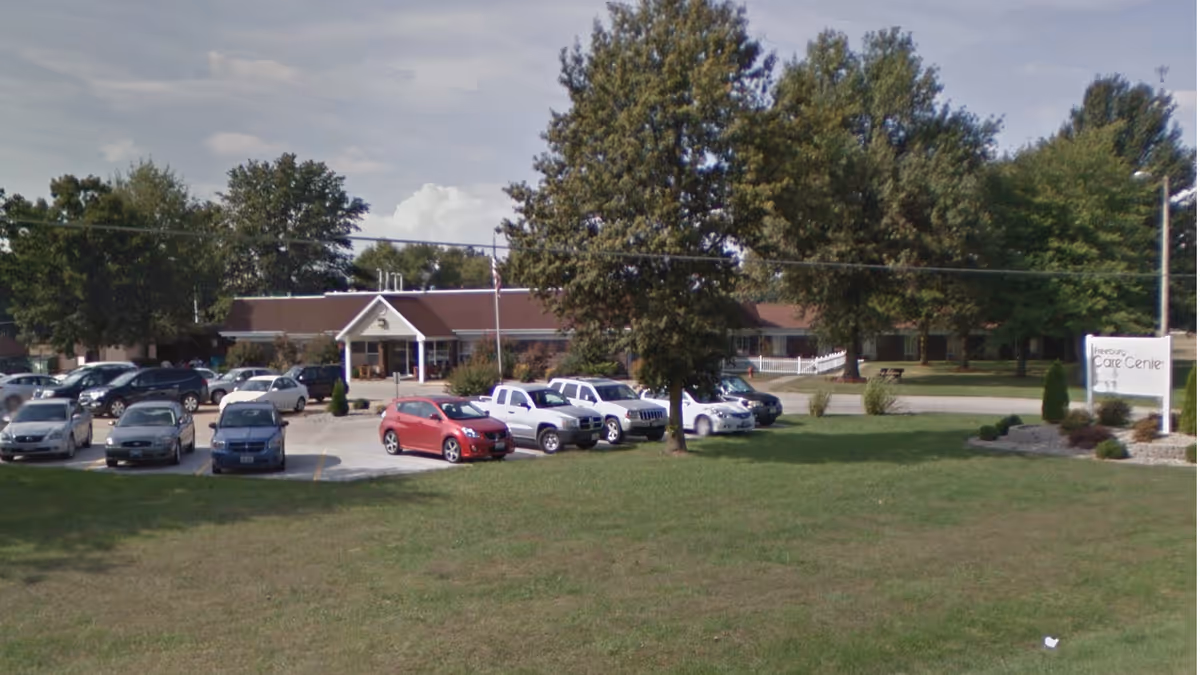 Front view of a single-story care center building with a parking lot, several cars, trees, and a grassy lawn.