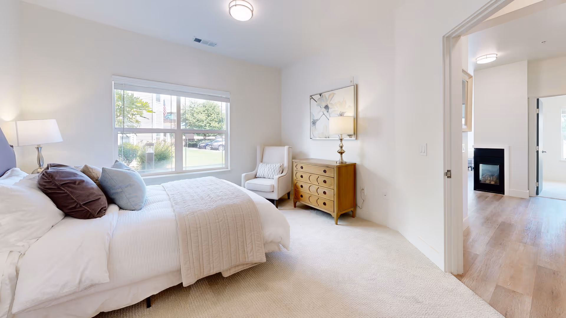 A bright and cozy bedroom with a large window showing greenery outside. The room features a neatly made bed with multiple pillows and a beige quilt, a wooden dresser with a lamp and artwork above it, and a comfortable armchair in the corner. The floor is carpeted, and an open doorway leads to a room with wooden flooring and a fireplace.
