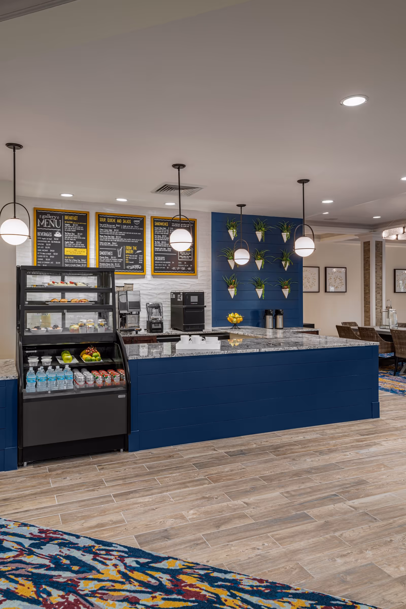 Interior view of a modern cafe or dining area with a blue counter, hanging globe pendant lights, a display case with bottled water, fruit, and desserts, and a menu board on the wall. The floor is wood-patterned tile and there is a colorful carpet in the foreground. The background shows a seating area with wicker chairs and framed artwork on the walls.