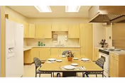 Small institutional kitchen and dining area with a table set for four, light wood cabinets, refrigerator, and stove.