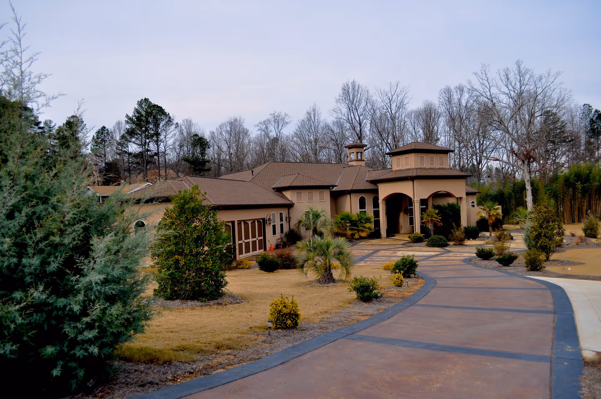 A wide driveway leads to the entrance of a single-story senior living facility building surrounded by landscaped bushes, small trees, and grass. The building has a tan exterior with a brown roof and features an arched covered entrance. Leafless trees are visible in the background under a cloudy sky.
