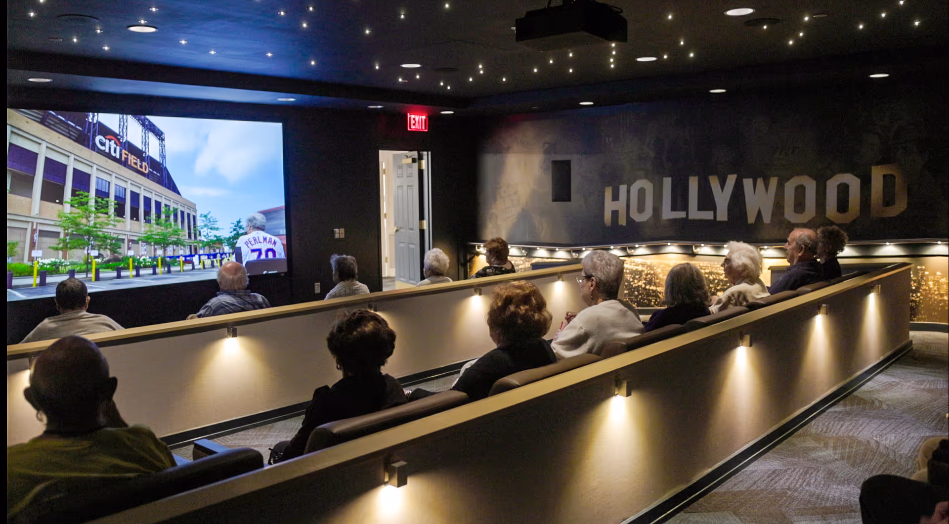 A group of elderly people seated in a dimly lit theater-style room watching a large screen showing an image of Citi Field stadium. The room has a Hollywood-themed wall decoration and soft lighting along the walls.