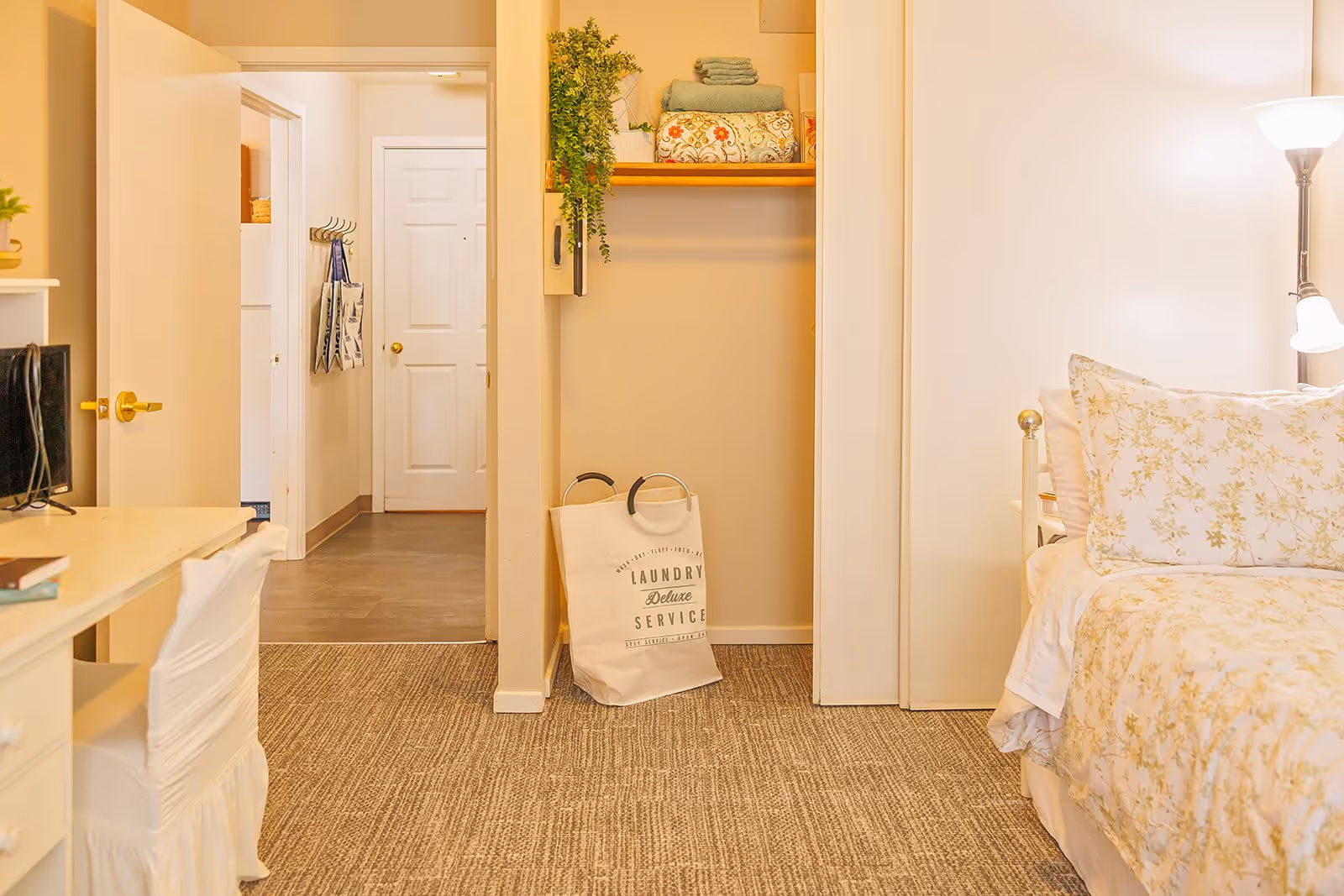 A cozy bedroom with a floral-patterned bedspread and pillows on the right, a white desk with a chair on the left, and an open closet in the center containing folded towels and a laundry bag. A hallway with white doors is visible in the background.