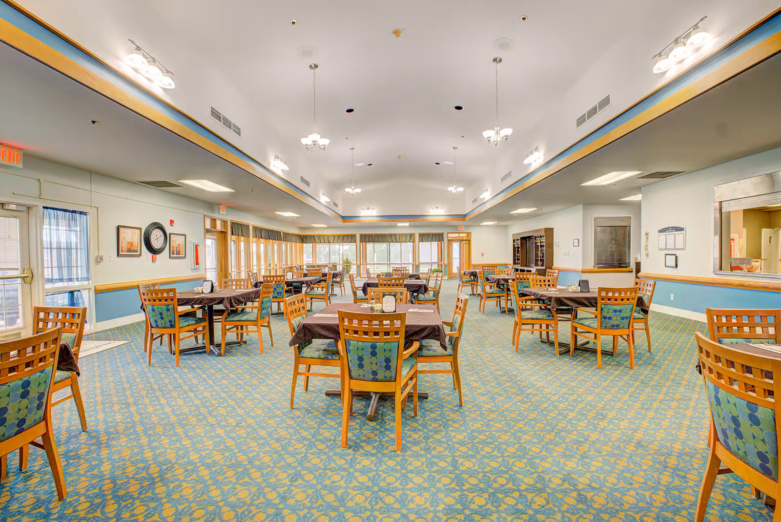 Spacious dining room with multiple wooden tables and chairs arranged neatly. The chairs have blue and green patterned cushions. The room has a blue and yellow patterned carpet, large windows along the far wall letting in natural light, and several ceiling light fixtures. There is a serving window on the right side and exit doors on the left and far end of the room.