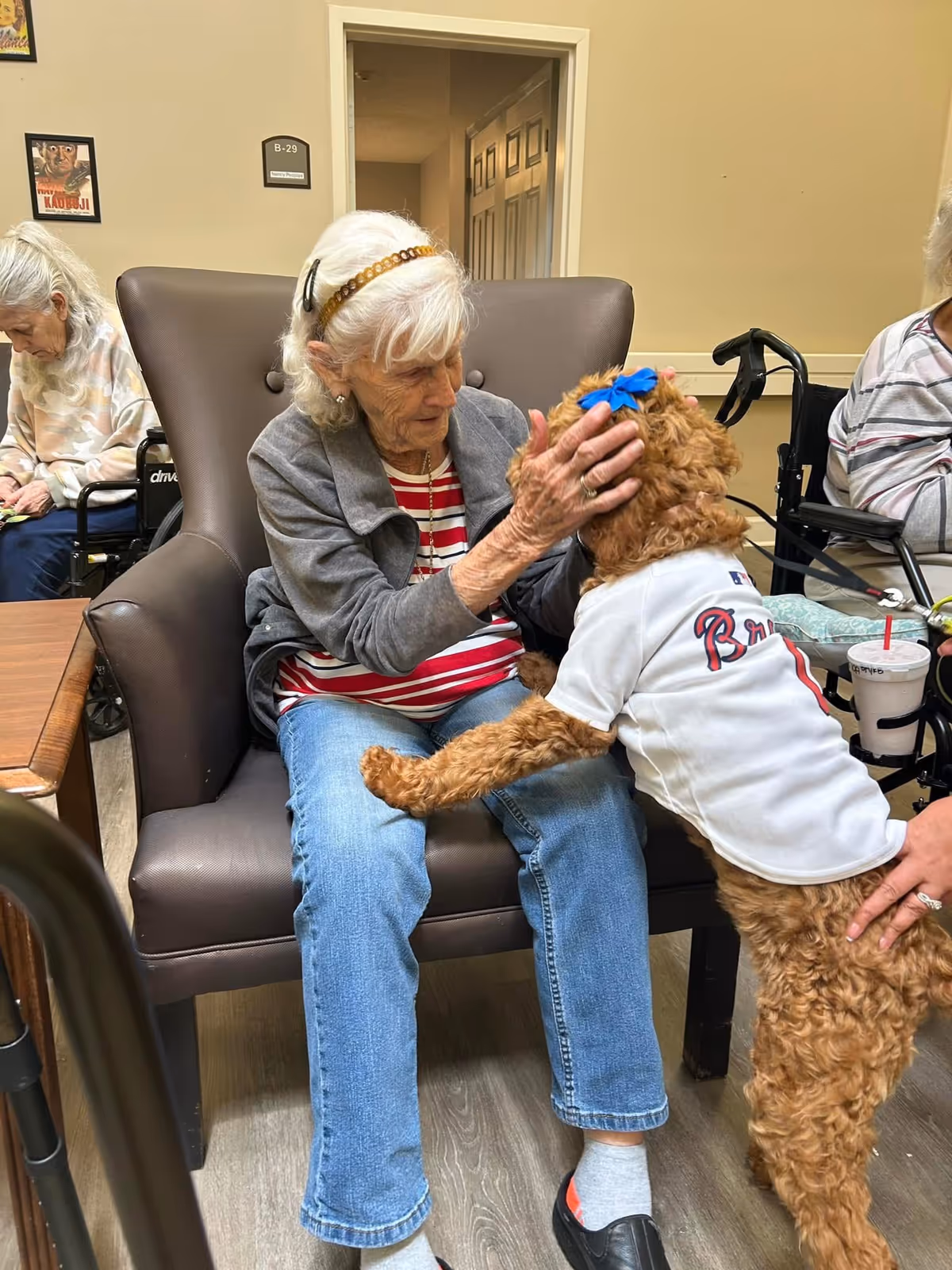 An elderly woman sitting in a brown armchair gently pets a small brown dog wearing a white jersey with 'Braves' written on it and a blue bow on its head. Two other elderly women are seated nearby, one in a wheelchair. The setting appears to be a common area in a senior living facility.
