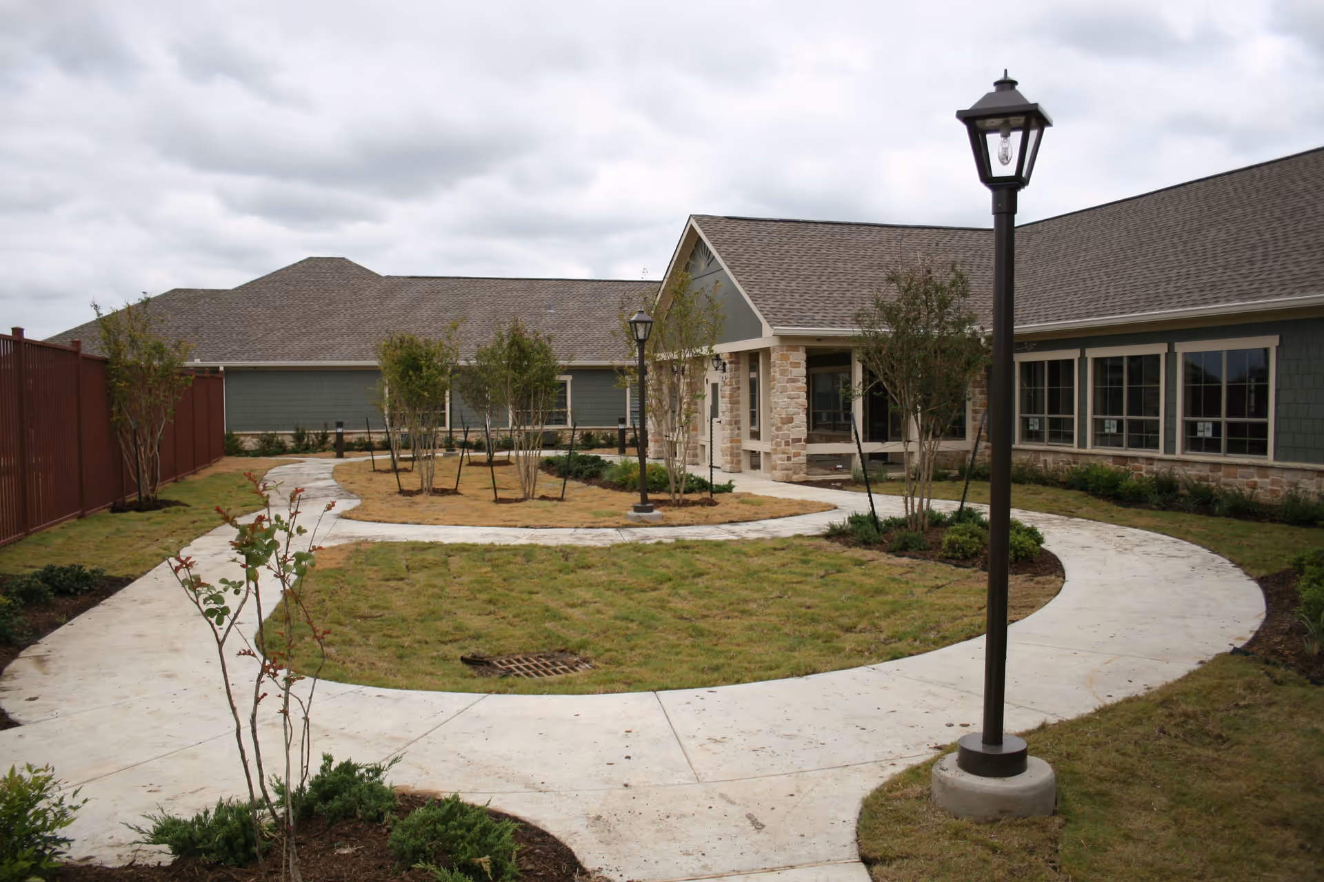 Outdoor courtyard area of a senior living facility with a curved concrete walkway, small trees, grass, and lamp posts. The building has green siding with stone accents and multiple windows under a cloudy sky.