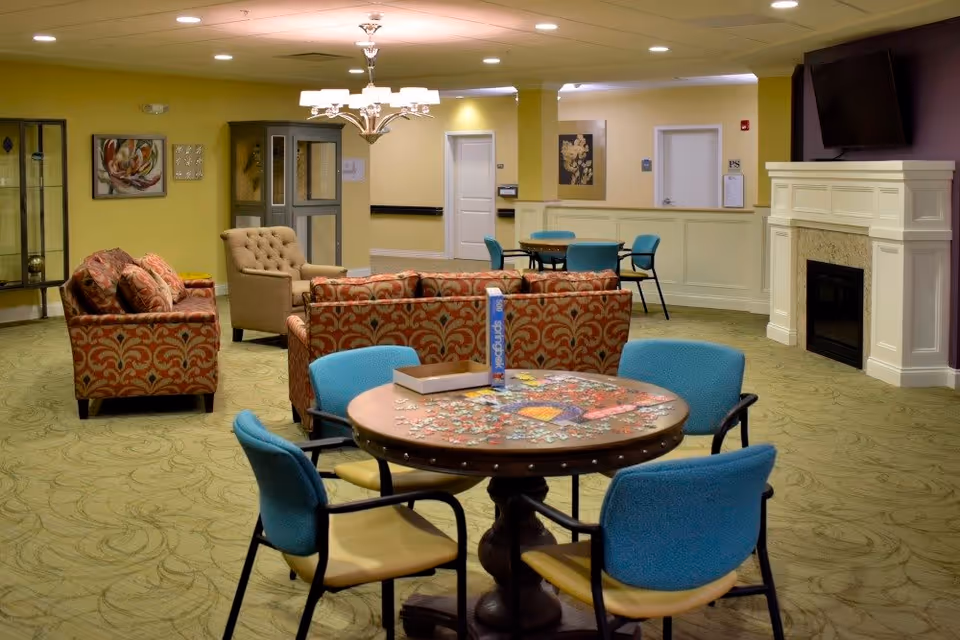 A cozy common area in a senior living facility with a round table in the foreground covered with a partially completed jigsaw puzzle and four blue chairs around it. In the background, there are patterned sofas and armchairs arranged near a white fireplace with a mounted TV above it. The walls are painted yellow and decorated with framed artwork, and the floor is carpeted with a green patterned design.