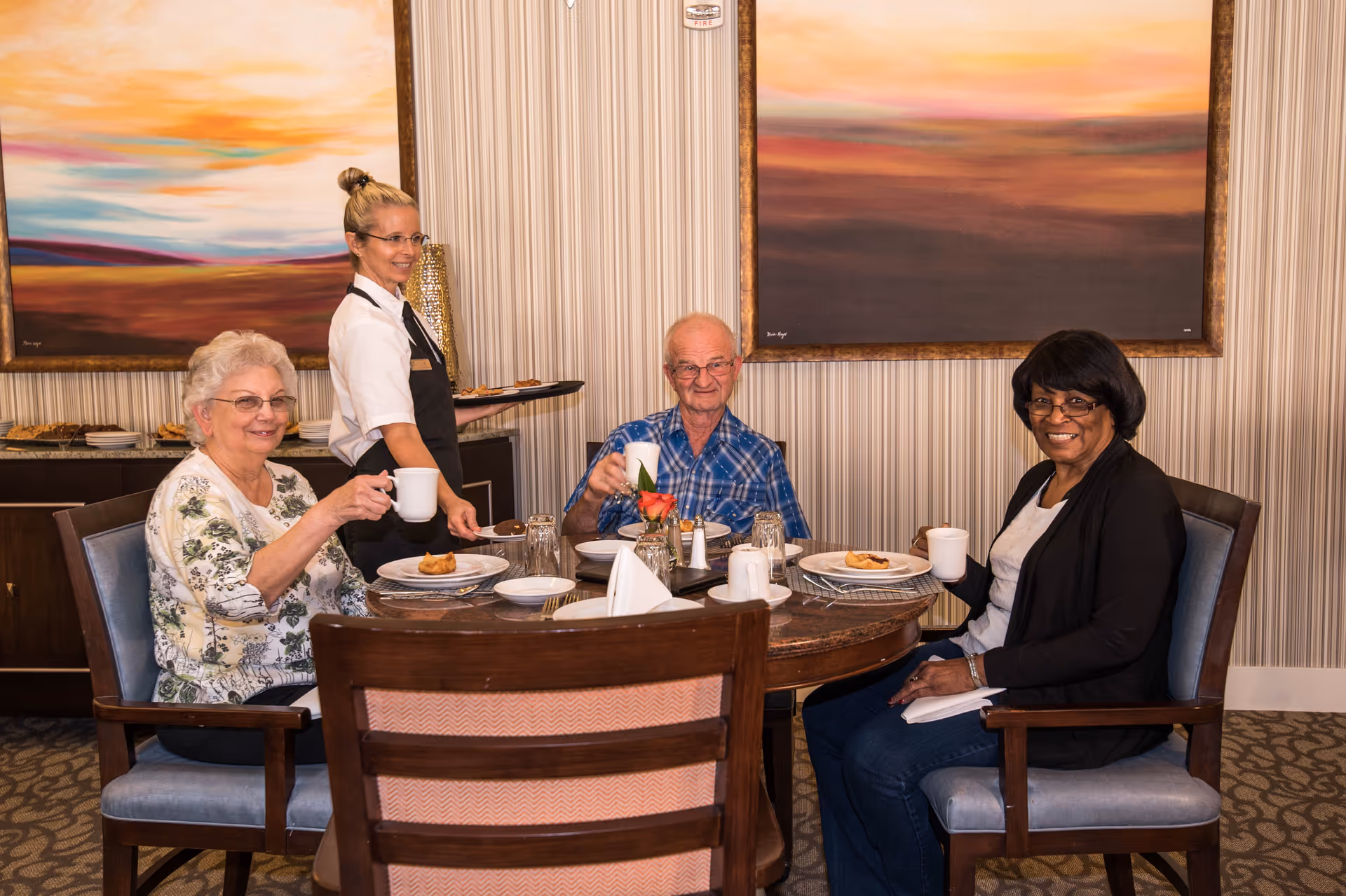 Three elderly people sitting around a dining table holding white mugs, smiling, with a waitress standing behind them holding a tray. The room has patterned wallpaper and two large landscape paintings on the wall.