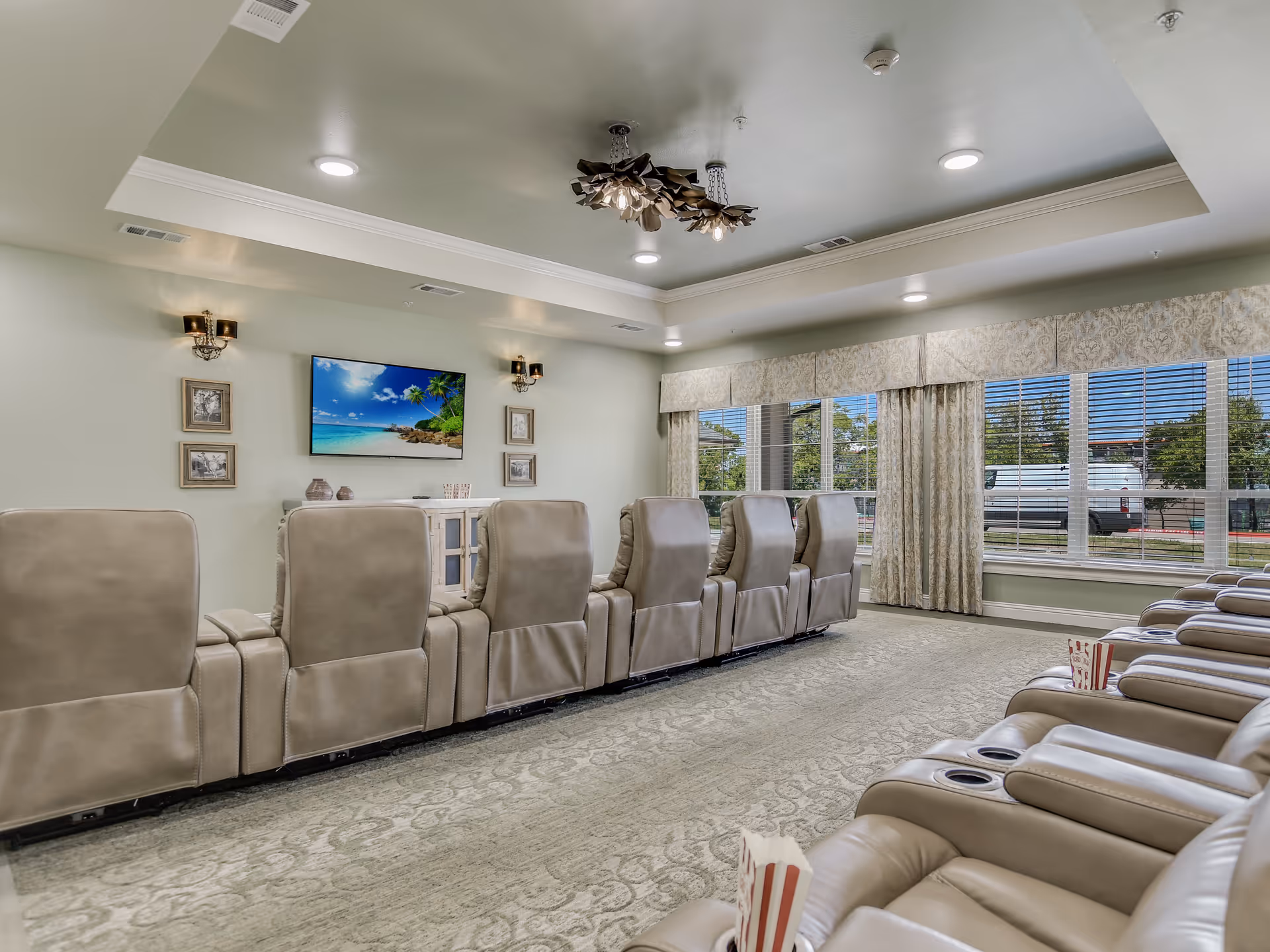 A senior living community media room with two rows of beige reclining theater seats facing a wall-mounted TV and large windows.