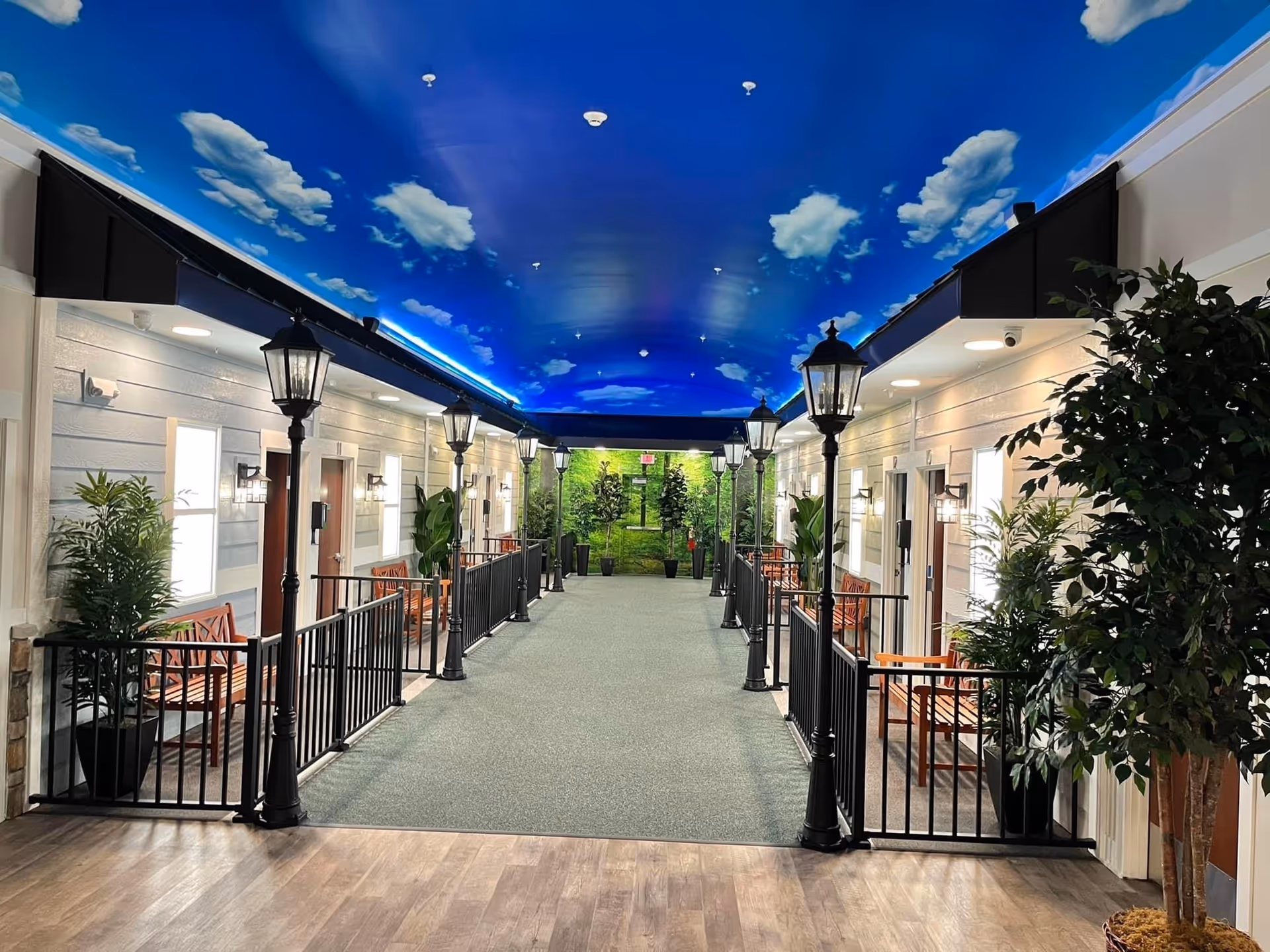 Indoor hallway designed to resemble an outdoor street with a blue ceiling painted with clouds, black street lamps lining both sides, wooden benches, potted plants, and doors leading to rooms on either side.