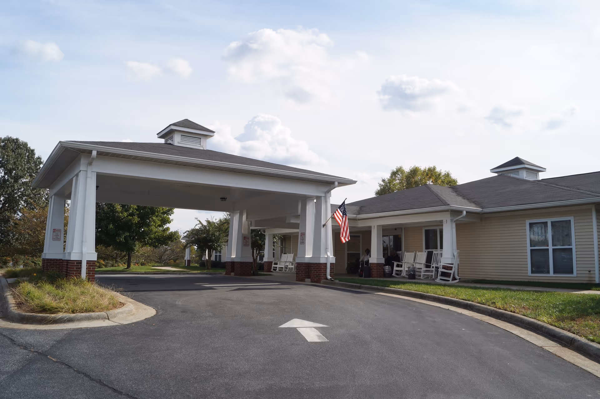 Front entrance of a senior living facility with a covered porte-cochere, rocking chairs on the porch, and an American flag.