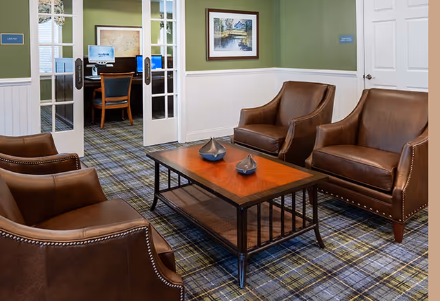 A cozy sitting area with four brown leather armchairs arranged around a wooden coffee table with decorative items on it. The room has green walls with white wainscoting and a plaid carpet. In the background, there is a room with computer desks and chairs visible through glass double doors.