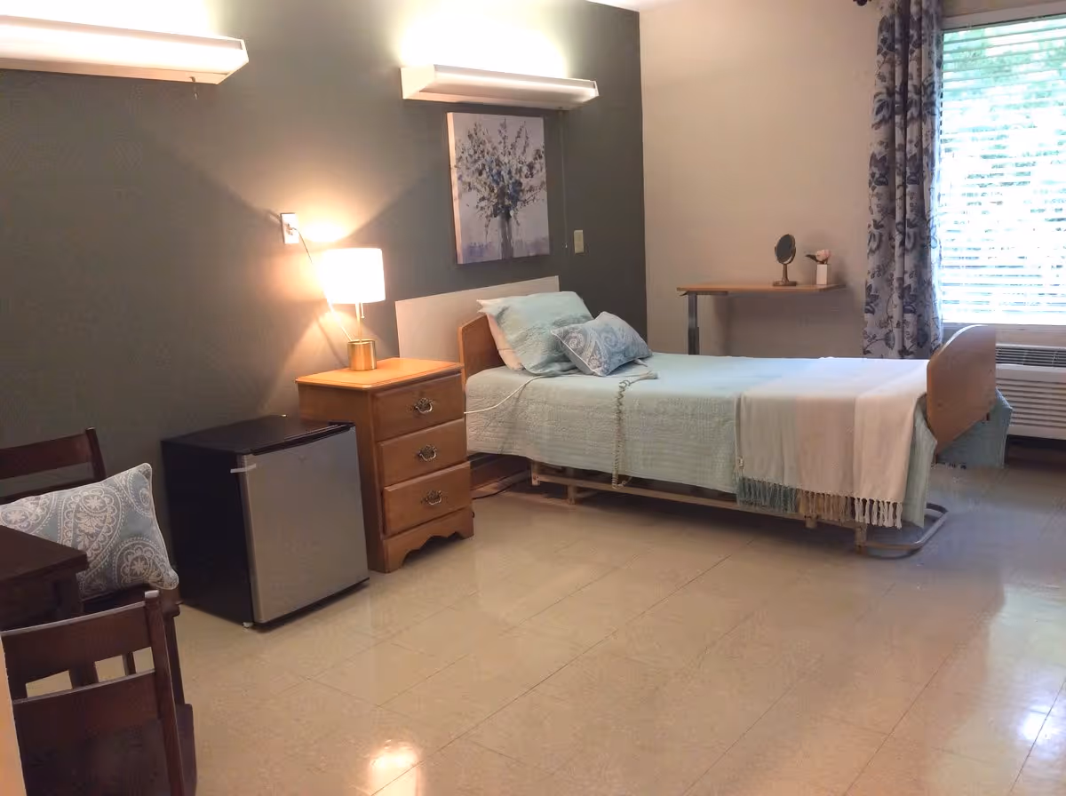 A simple, clean bedroom in a senior living facility with a single bed made with light blue bedding and a white throw blanket. Next to the bed is a wooden nightstand with a lamp and a small refrigerator underneath. There is a chair with a patterned cushion, a small table with a mirror and a flower vase, and a window with floral curtains letting in natural light.