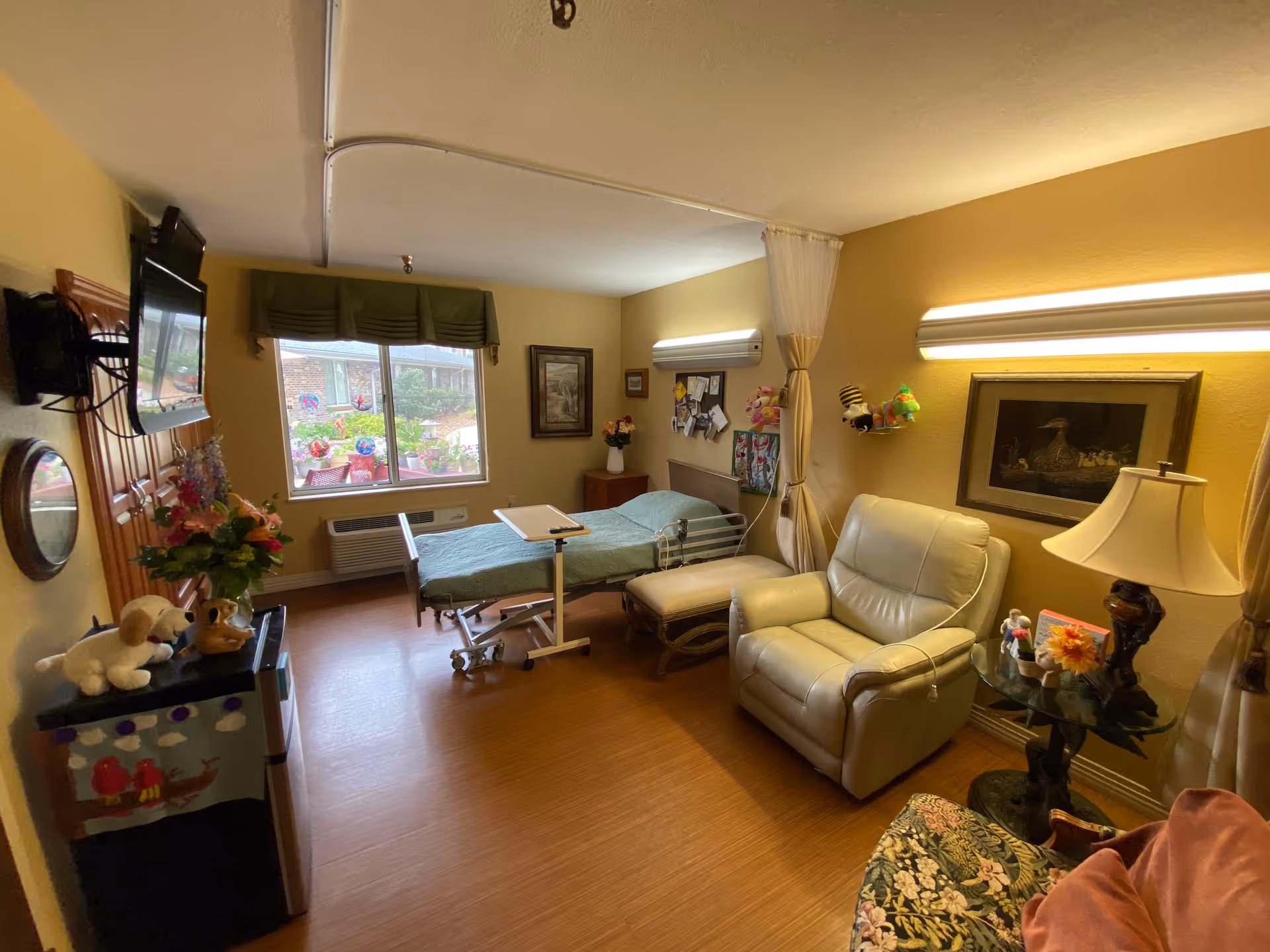 A cozy room in a senior living facility with a hospital bed, a reclining chair, a small table with a lamp, and a window showing a garden outside. The walls are painted yellow, and there are various decorations including framed pictures, stuffed animals, and flowers.