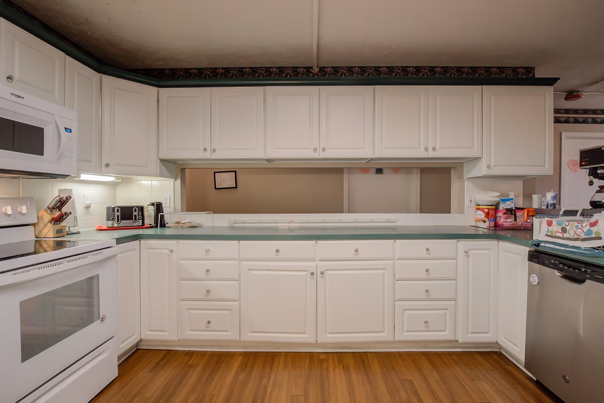 A clean kitchen with white cabinets and drawers, green countertops, a white stove with a microwave above it, a toaster, a knife block, and a dishwasher. The floor is wooden, and there is a pass-through window above the counter.