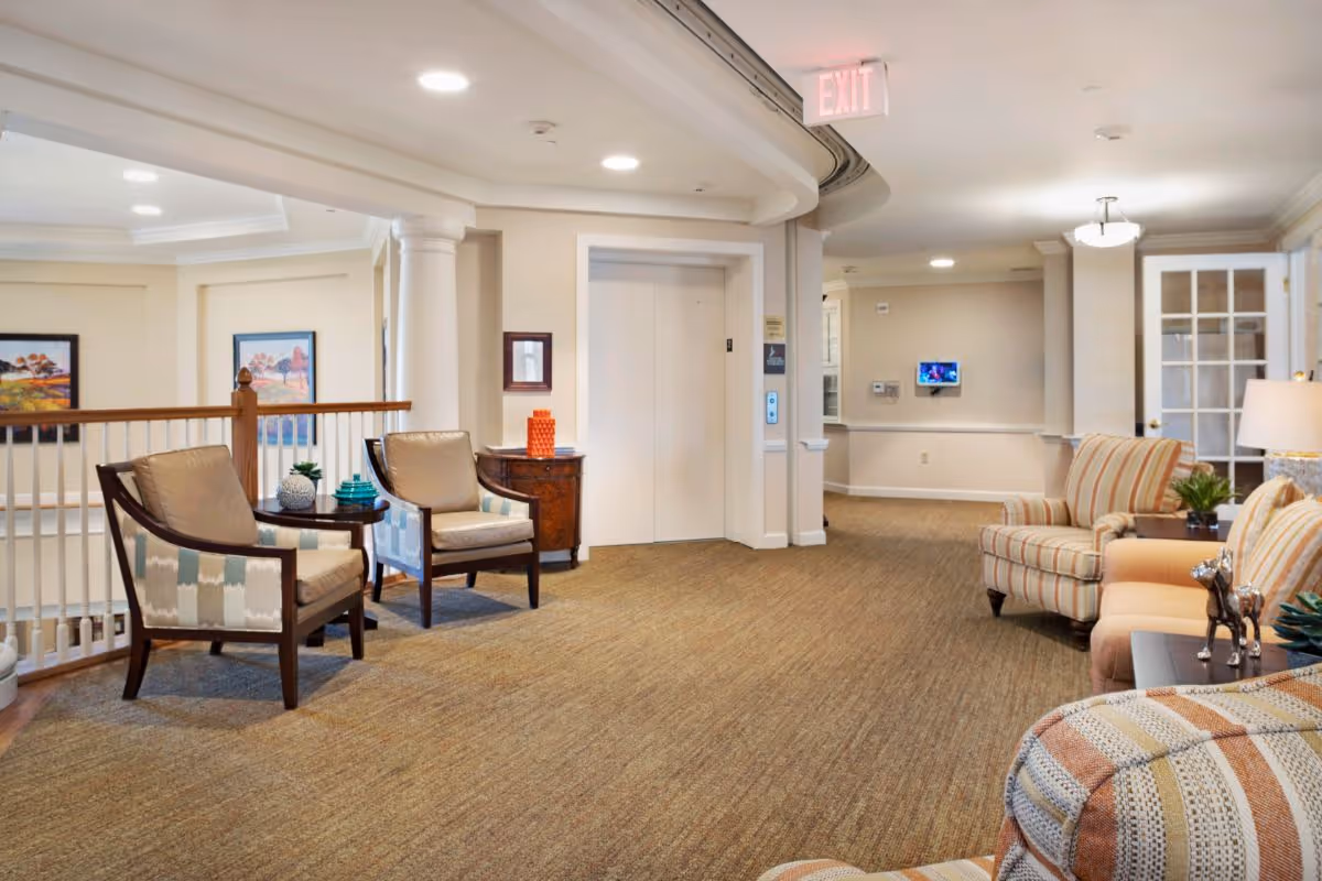 A well-lit interior common area in a senior living facility featuring comfortable seating including two patterned armchairs with a small round table between them, a striped sofa, and an armchair. There is an elevator with closed doors in the background, a small wooden cabinet with decorative items, framed artwork on the walls, and a carpeted floor. The space has neutral-colored walls and ceiling lights, with an exit sign above the elevator.