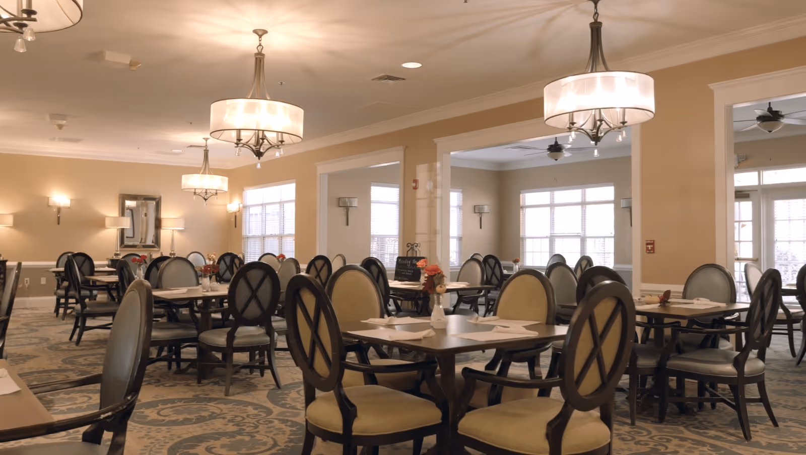 A spacious dining room with multiple tables and chairs arranged neatly. The room features large windows allowing natural light to fill the space, beige walls, elegant chandeliers hanging from the ceiling, and a patterned carpet. Each table is set with placemats and small flower vases.