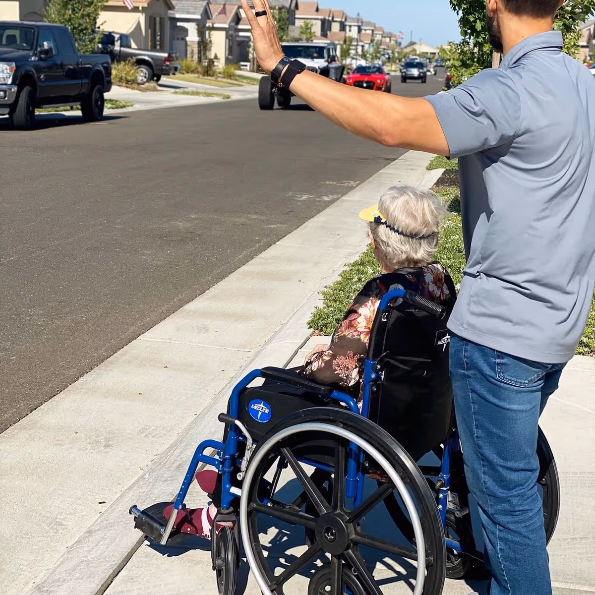 An elderly woman in a wheelchair is being assisted by a man standing beside her on a sidewalk in a residential neighborhood. The man is raising his hand, possibly signaling to oncoming traffic. Several parked cars and houses are visible in the background under a clear sky.