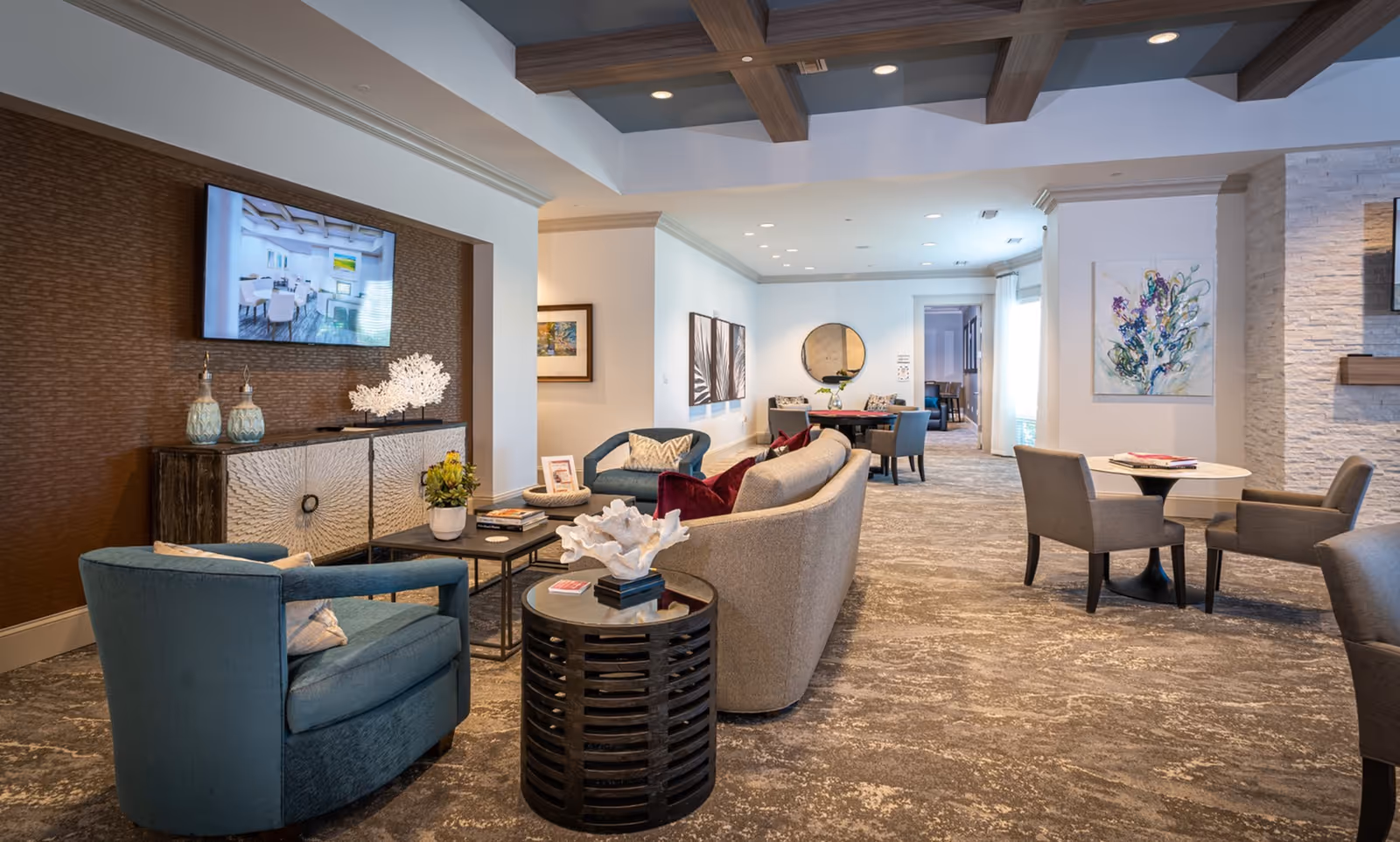 A spacious and well-lit living room area in a senior living facility with modern furniture including blue armchairs, a beige curved sofa with red cushions, and small round tables. The room features a mounted flat-screen TV, decorative artwork on the walls, and a carpeted floor with a textured pattern. The ceiling has wooden beams and recessed lighting.