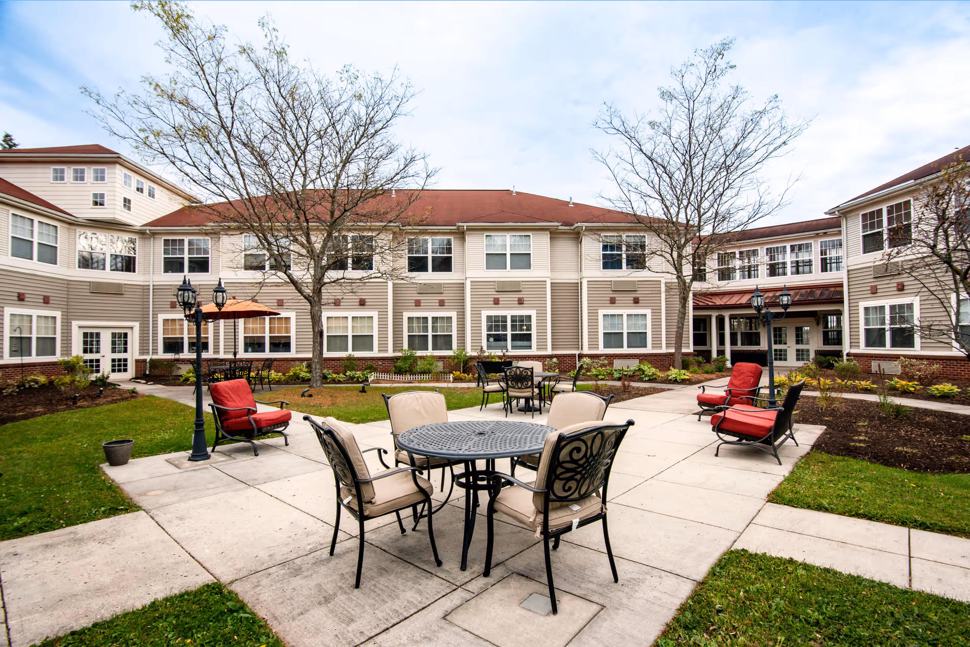 Outdoor courtyard area of a senior living facility with metal tables and cushioned chairs arranged on a paved patio. The courtyard is surrounded by a two-story beige building with many windows. There are leafless trees and green grass around the patio, and lamp posts with lantern-style lights.
