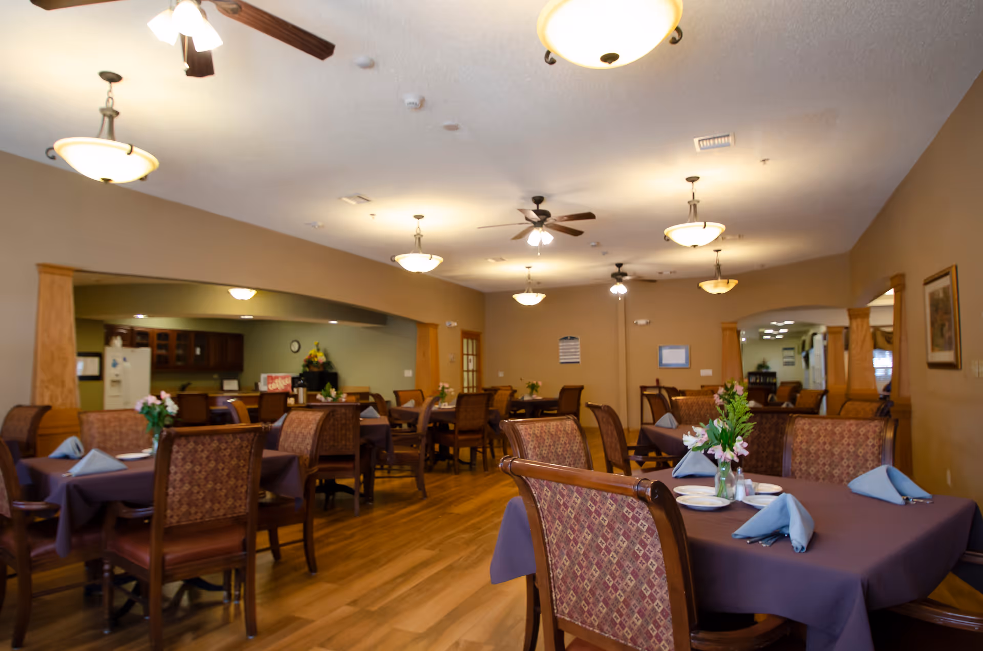Interior view of a dining room in a senior living facility with multiple tables covered in purple tablecloths, each set with plates, napkins, and floral centerpieces. The room features wooden chairs with patterned upholstery, ceiling fans, and warm lighting fixtures. In the background, there is a kitchen area with cabinets and a refrigerator.