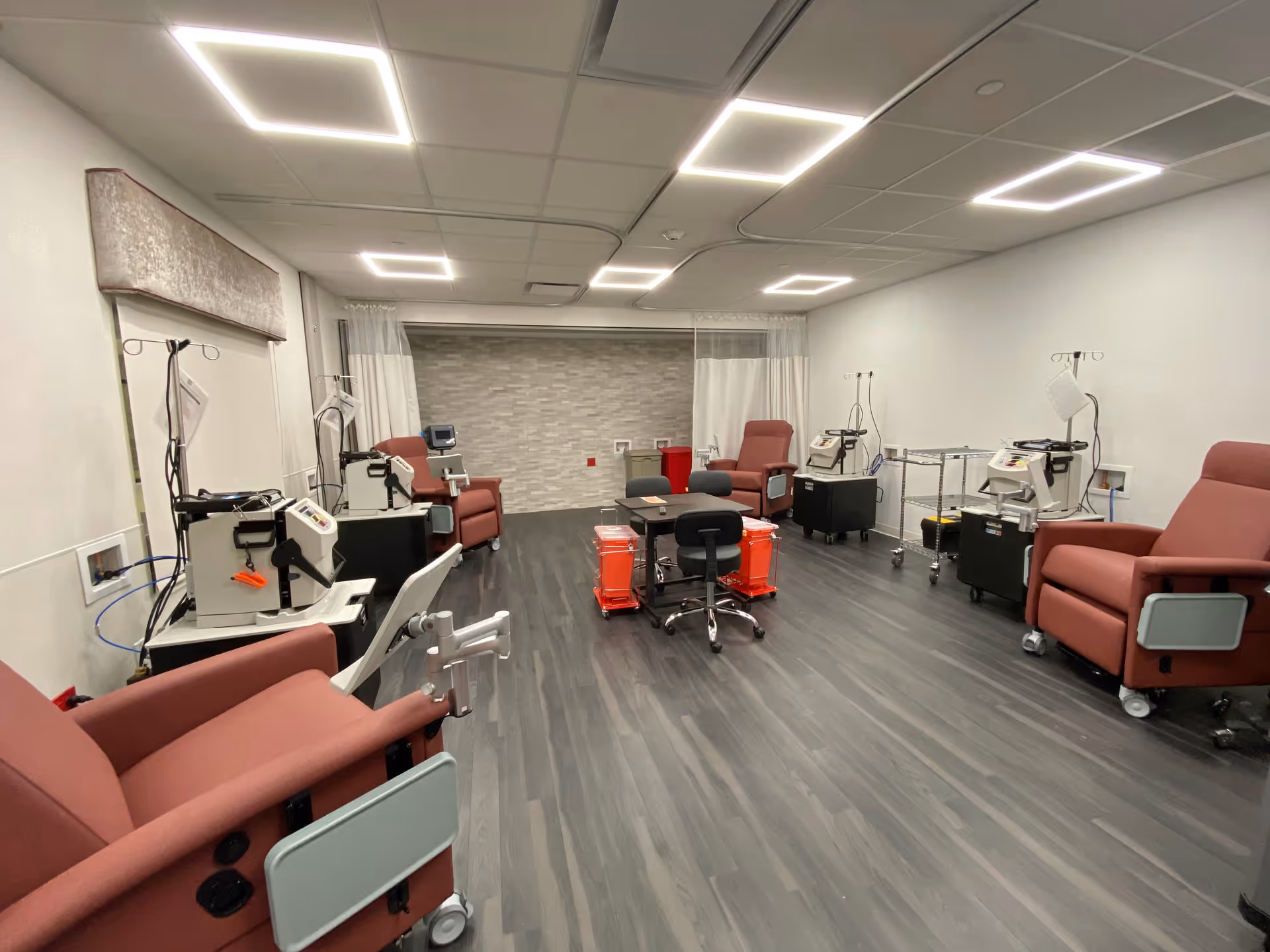 A medical treatment room with several maroon recliner chairs spaced around the room, each equipped with medical devices and IV stands. The room has a modern design with dark wood flooring, white walls, and bright square ceiling lights. There are white curtains along the back wall and medical carts with supplies in the center.